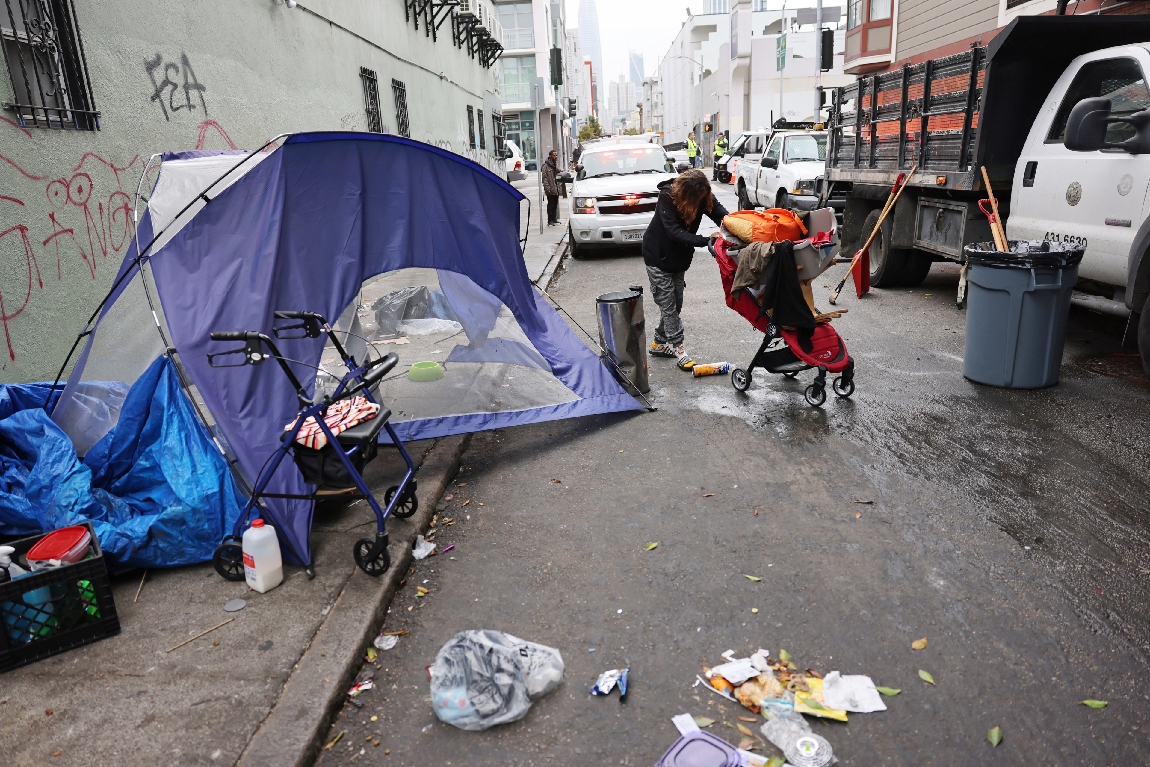 A street scene shows a makeshift tent, a stroller, and scattered items. A person is organizing belongings while city workers in trucks are nearby.