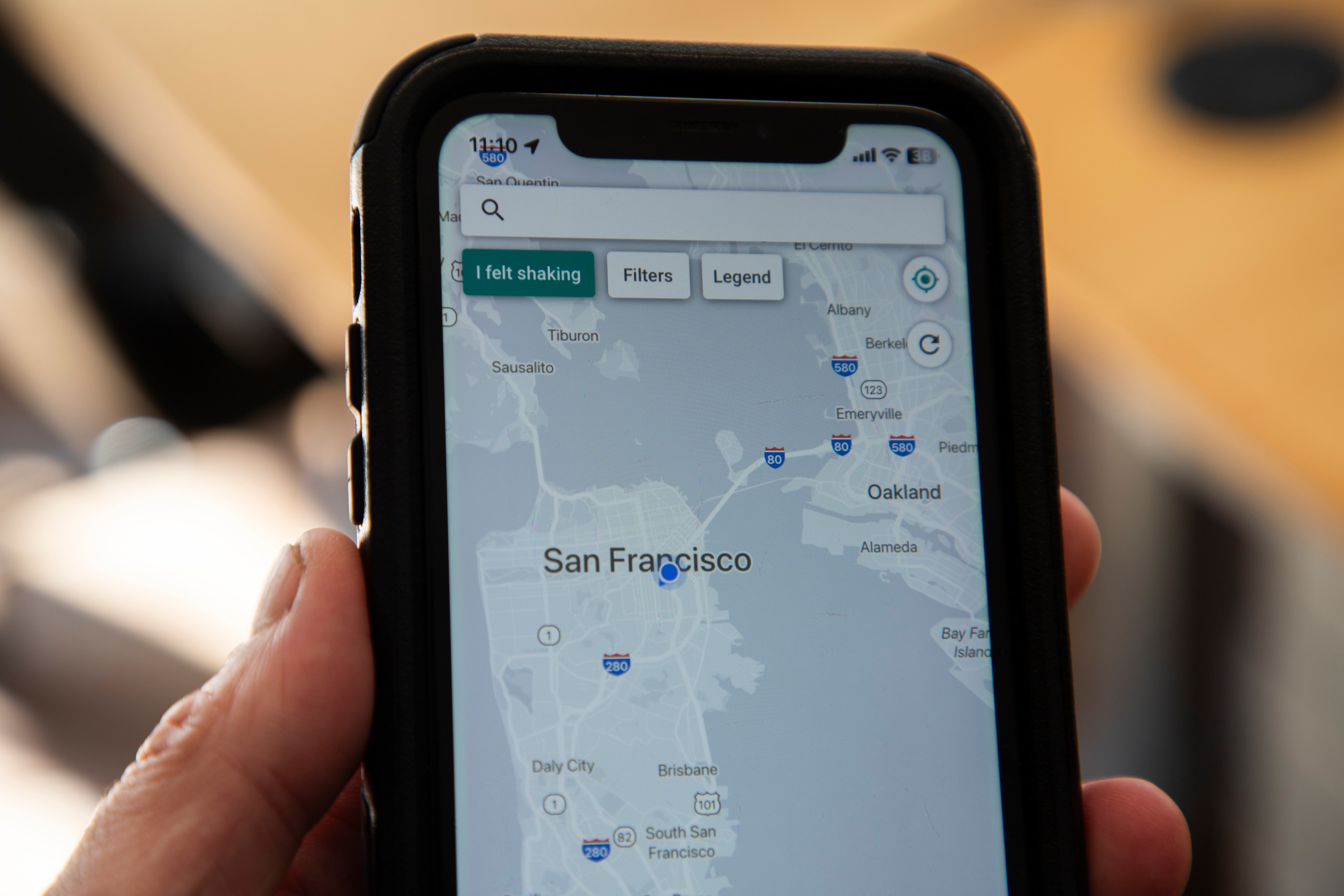 A hand holds a smartphone displaying a map of San Francisco with a &quot;I felt shaking&quot; button on the screen, suggesting an earthquake app.