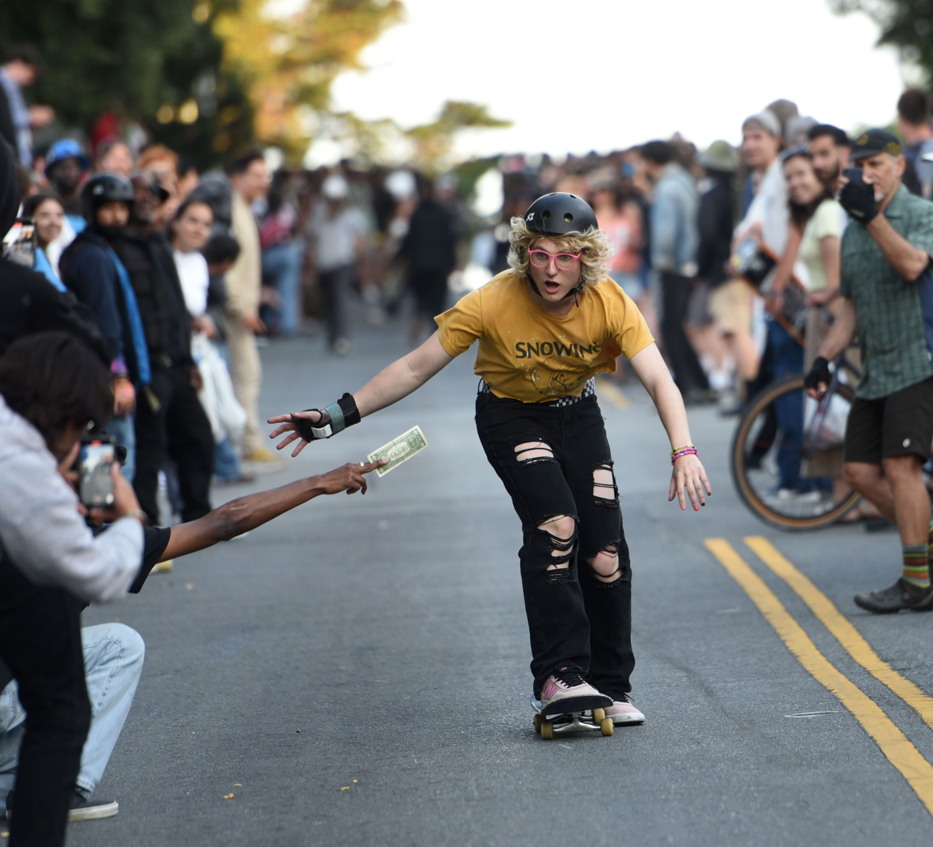 A skateboarder in a yellow shirt and helmet rides down a street, reaching for money held by a spectator. A crowd lines the road, watching intently.