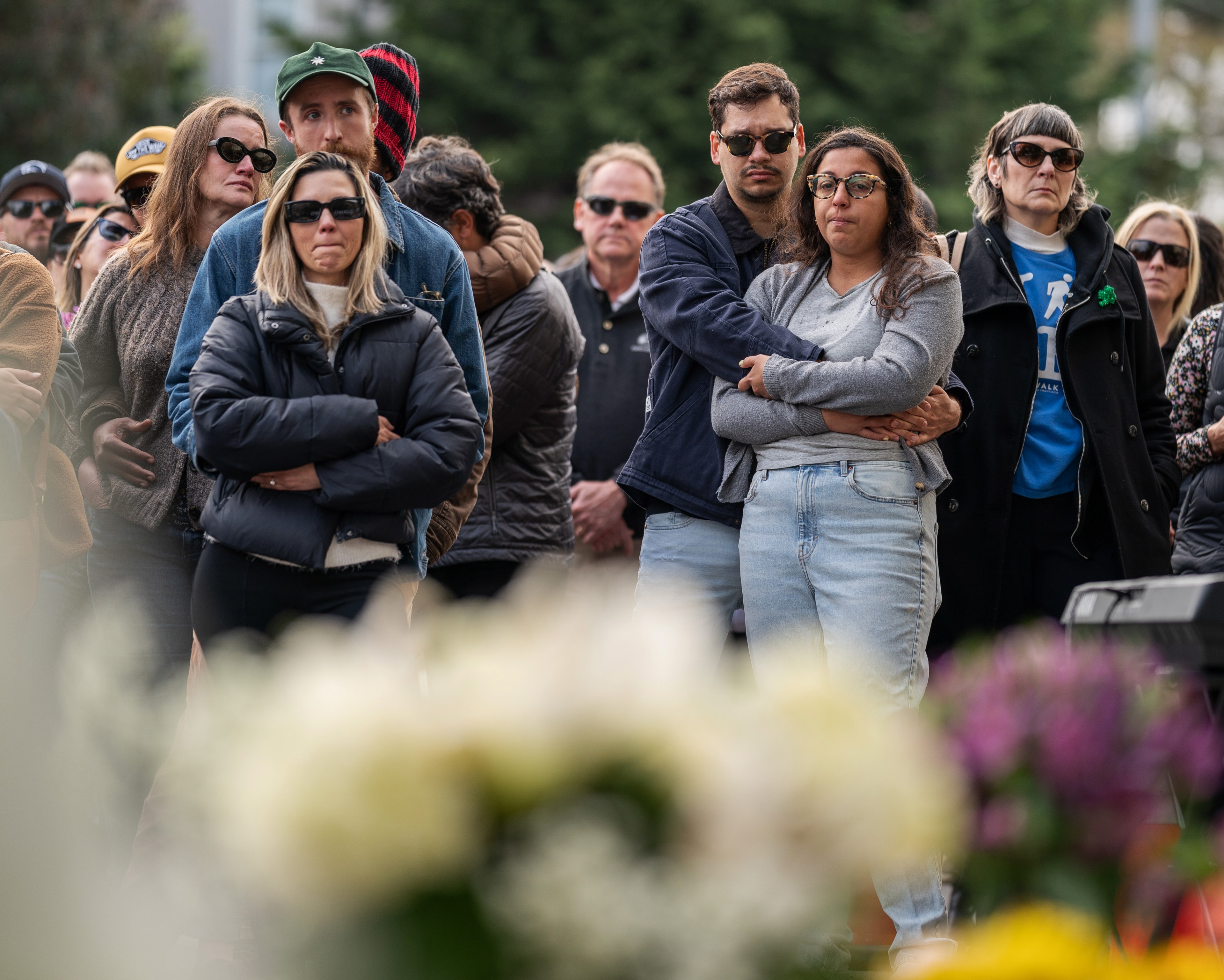 A group of people are gathered outdoors, looking somber. They are dressed in casual, warm clothing, wearing sunglasses. The foreground shows blurred flowers.