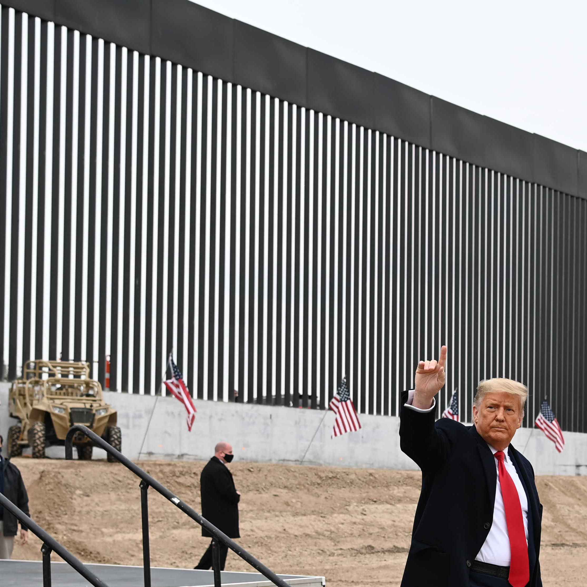 A man in a suit raises his hand near a tall border wall with vertical slats. Several American flags and a military-style vehicle are visible in the background.