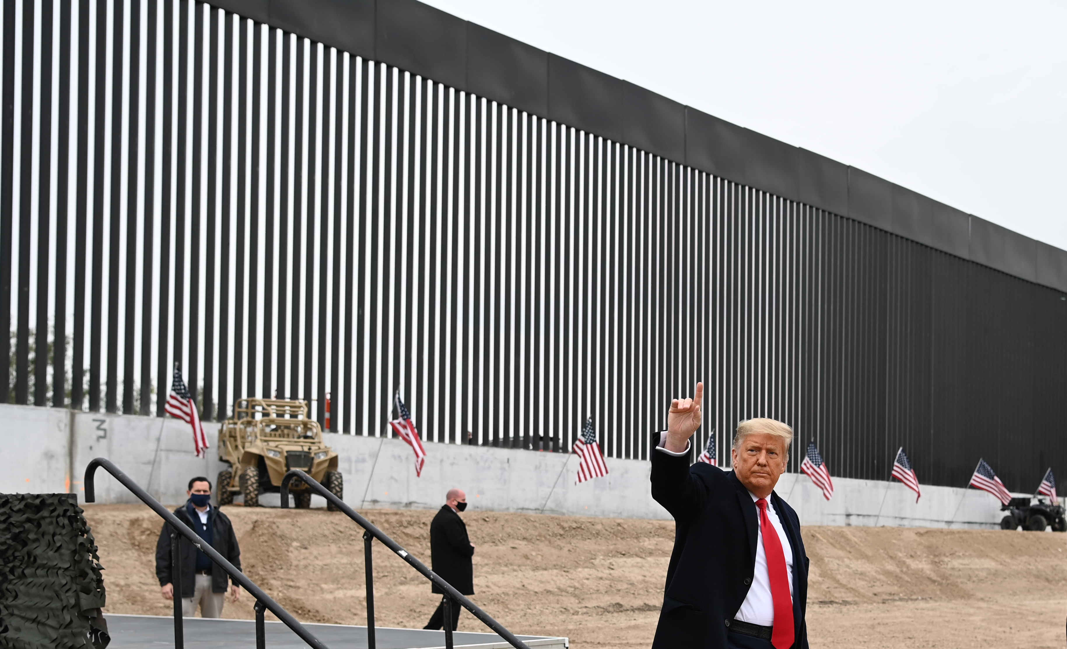 A man in a suit raises his hand near a tall border wall with vertical slats. Several American flags and a military-style vehicle are visible in the background.