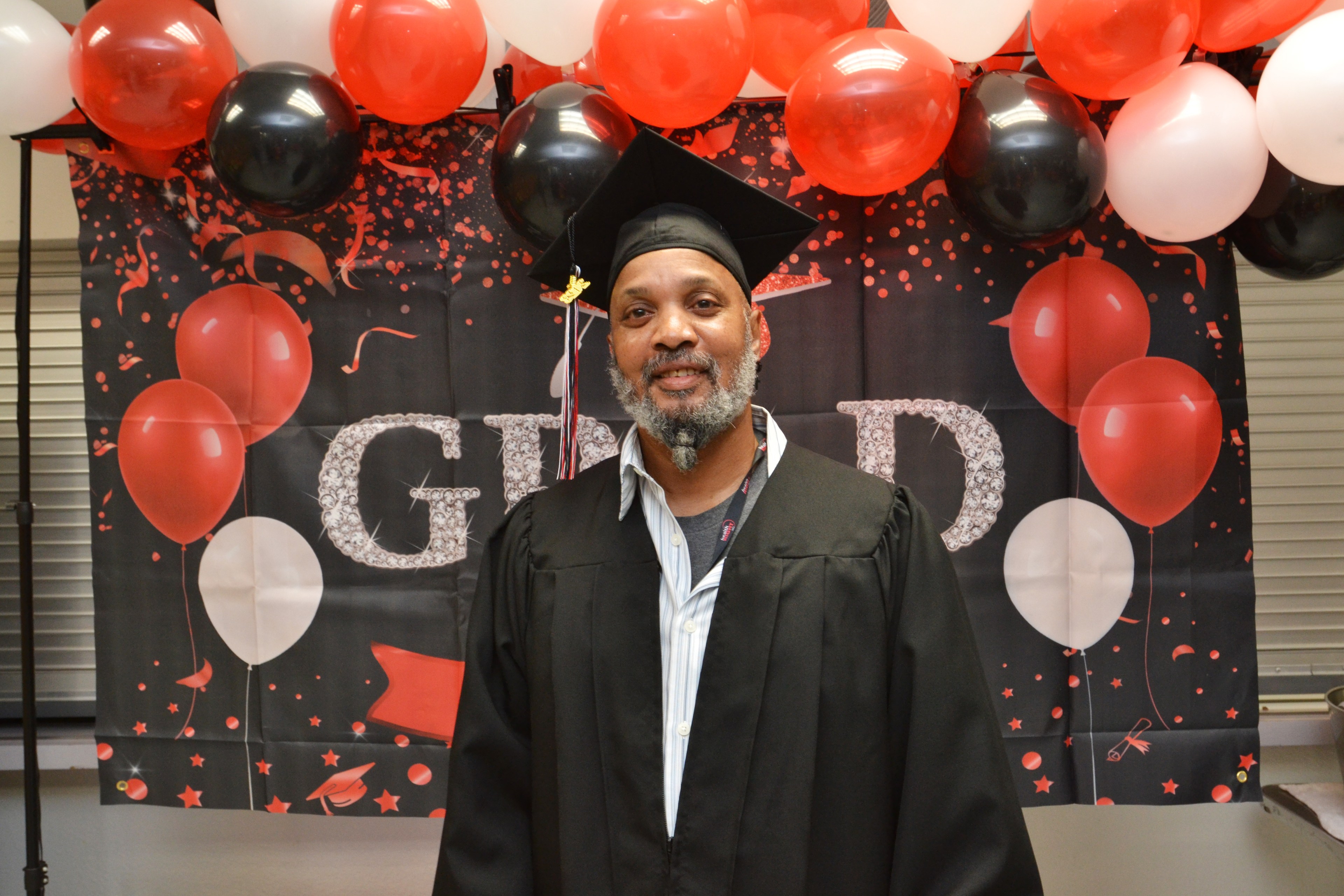 A man in a graduation gown and cap smiles in front of a festive backdrop with balloons. The backdrop features red, black, and white colors with celebratory designs.