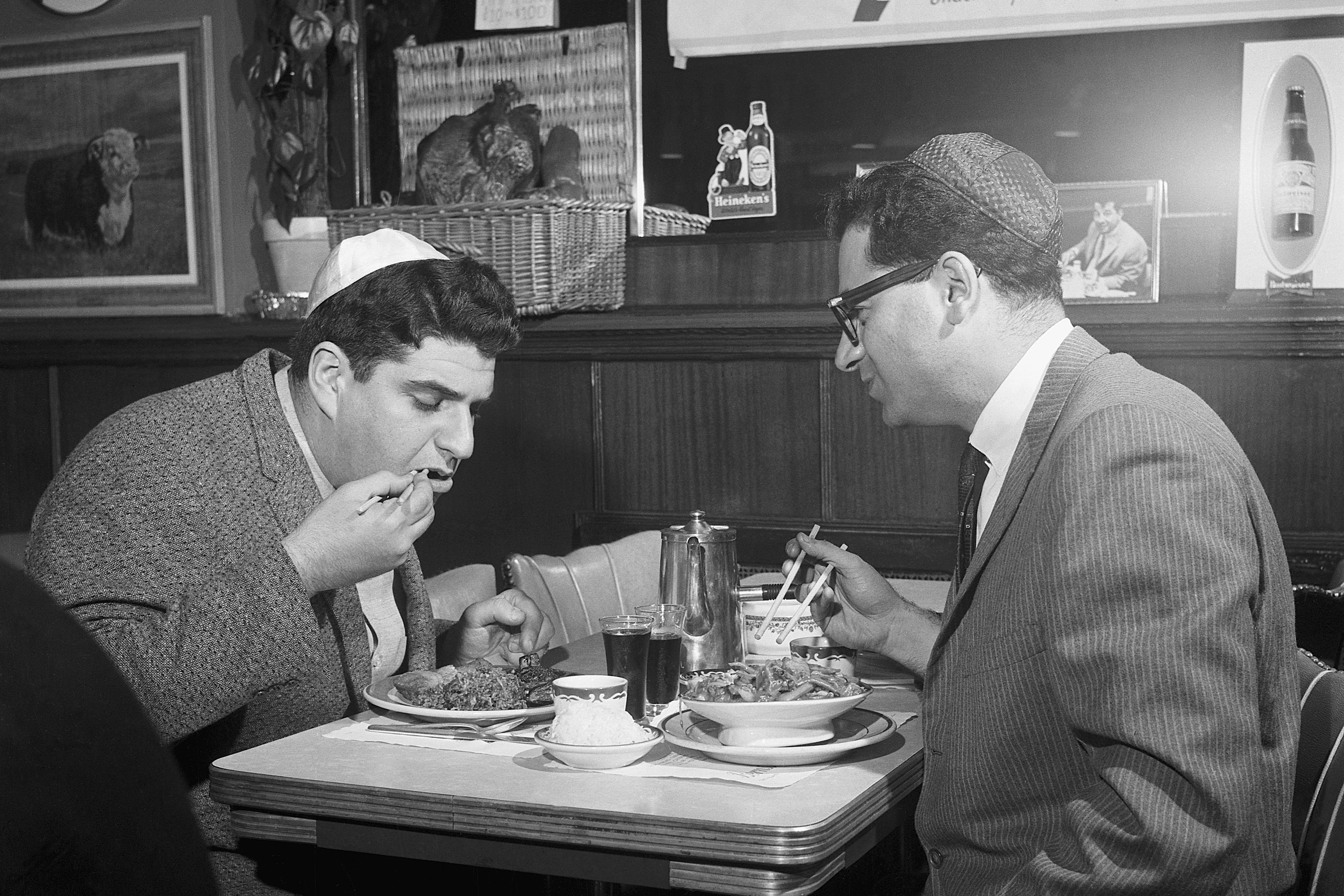 Two men wearing yarmulkes are eating at a table with Chinese food. A sign behind them reads "Saturday Chinese Night at Bernstein's on Essex St."