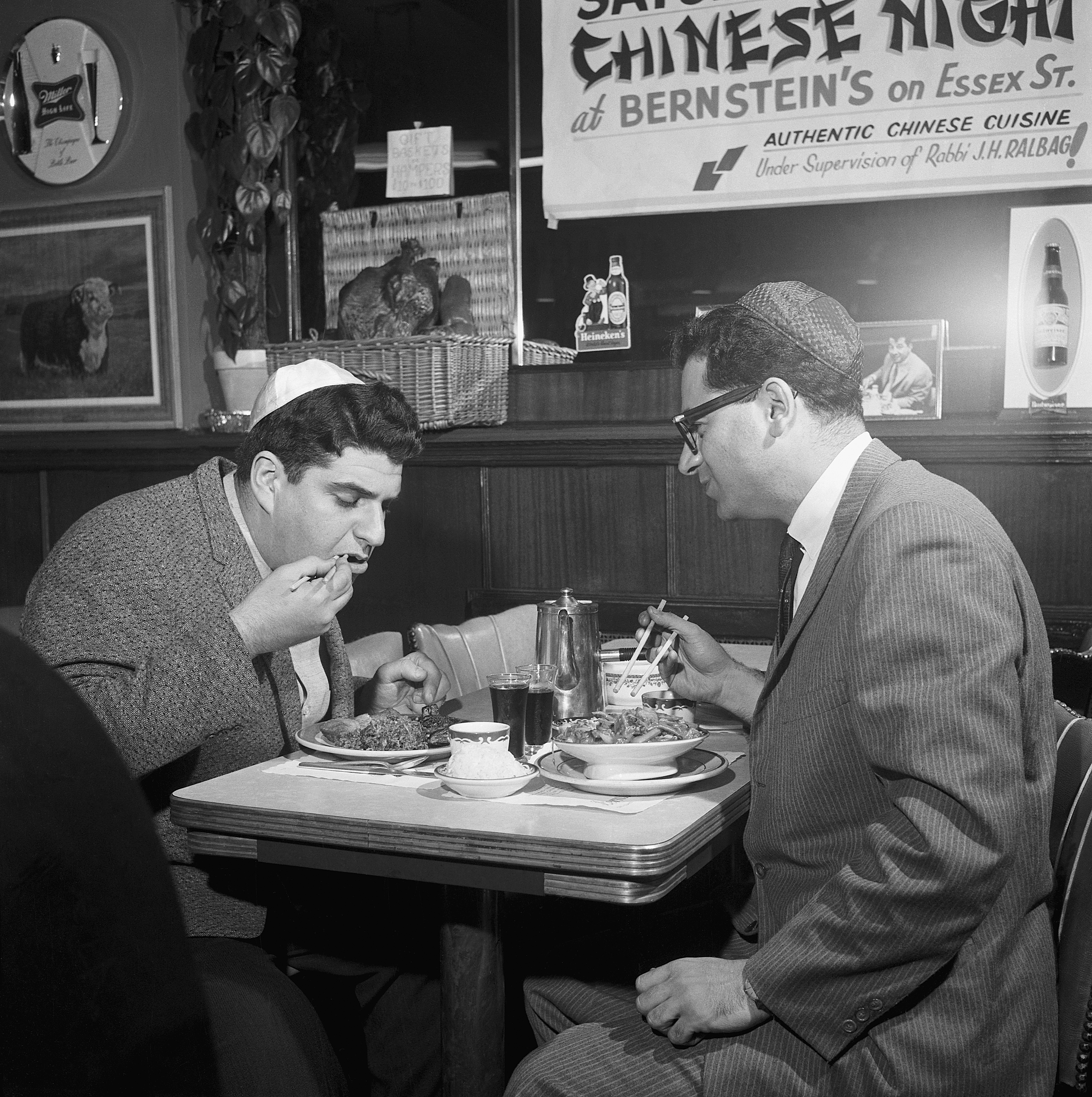 Two men wearing yarmulkes are eating at a table with Chinese food. A sign behind them reads "Saturday Chinese Night at Bernstein's on Essex St."