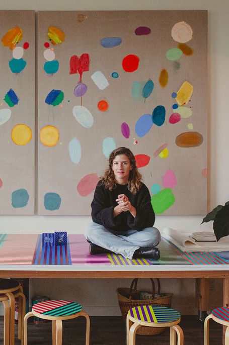 A person sits cross-legged on a colorful table with striped stools, framed by vibrant abstract art pieces and surrounded by lush potted plants.