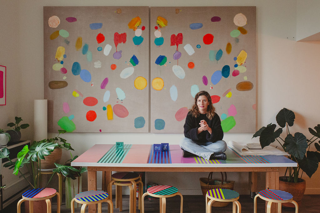 A person sits cross-legged on a colorful table with striped stools, framed by vibrant abstract art pieces and surrounded by lush potted plants.