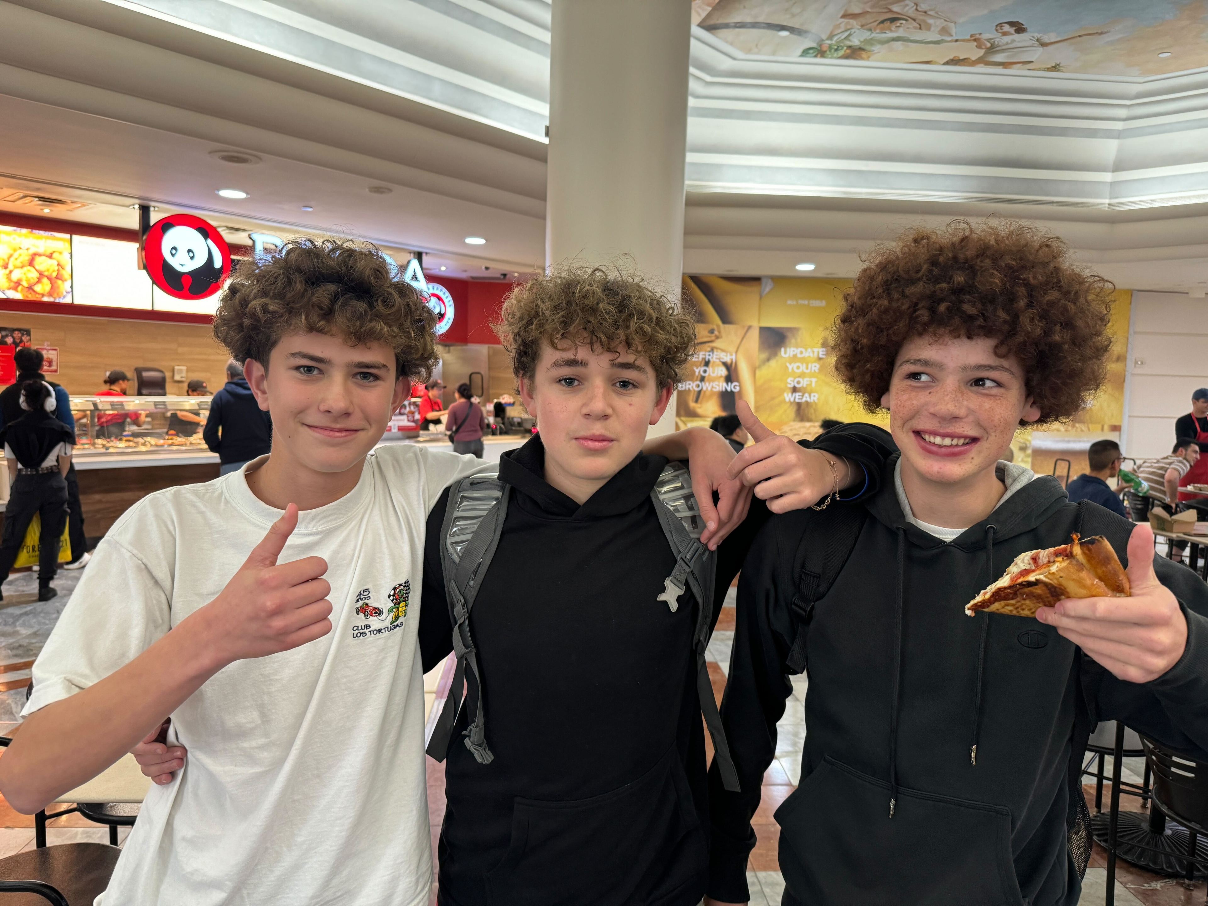 Three smiling young people stand in a food court, giving thumbs up. One holds a slice of pizza. They're in front of a Panda Express counter.