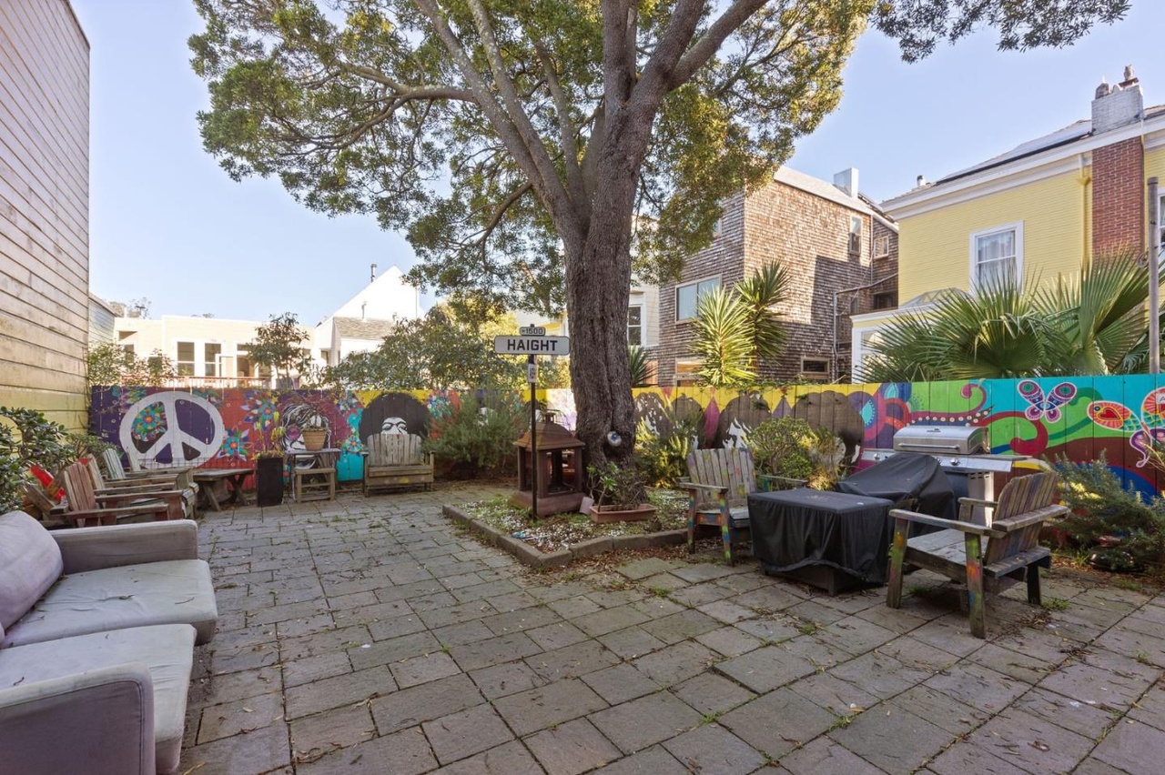 A colorful backyard features a large tree, a vibrant mural, wooden chairs, a sign saying &quot;Haight,&quot; and a barbecue grill, all on a stone patio.