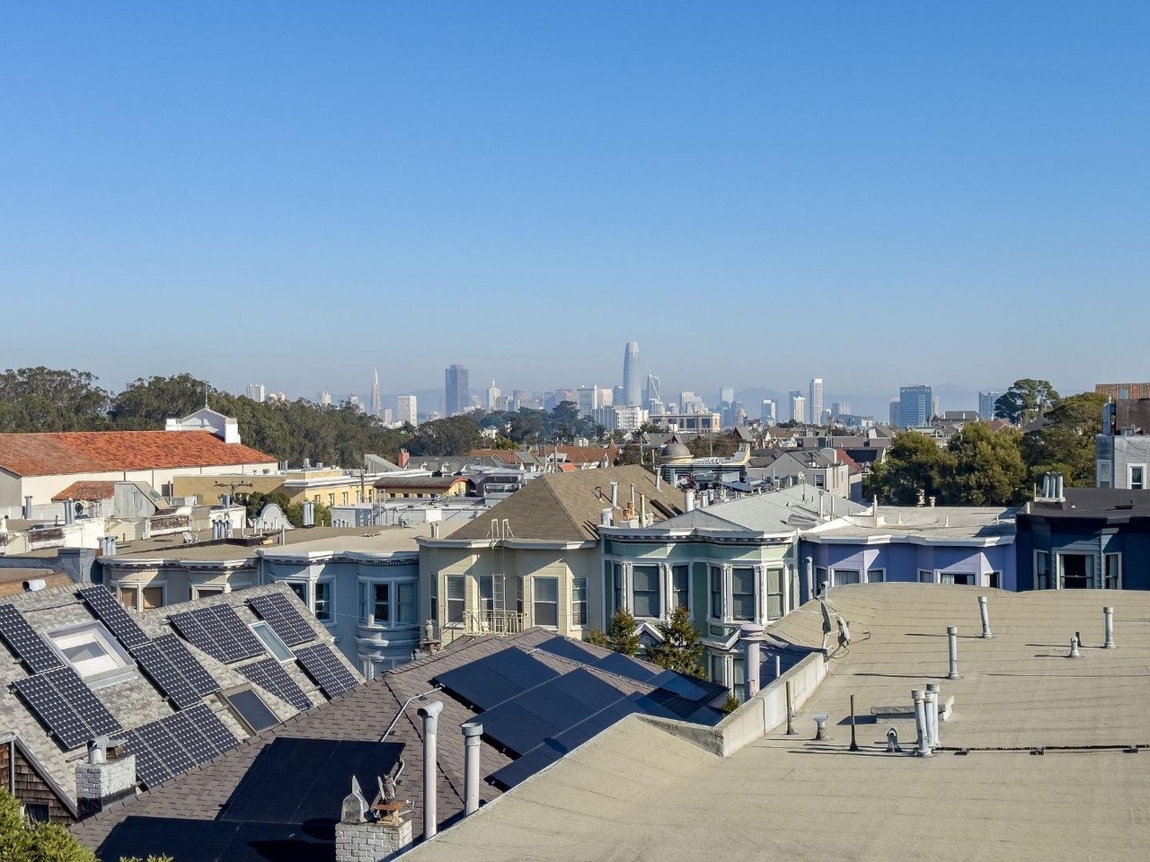 The image shows a cityscape with colorful houses in the foreground, several topped with solar panels, and a distant skyline under a clear, blue sky.