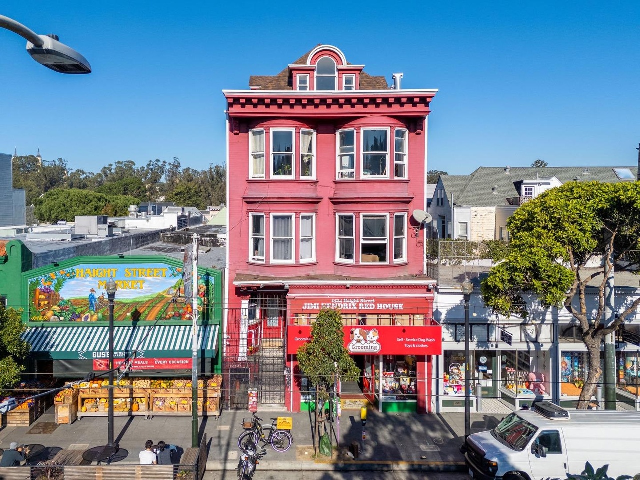 A vibrant pink building with bay windows stands between Haight Street Market and another shop, framed by trees and a parked van on a sunny day.