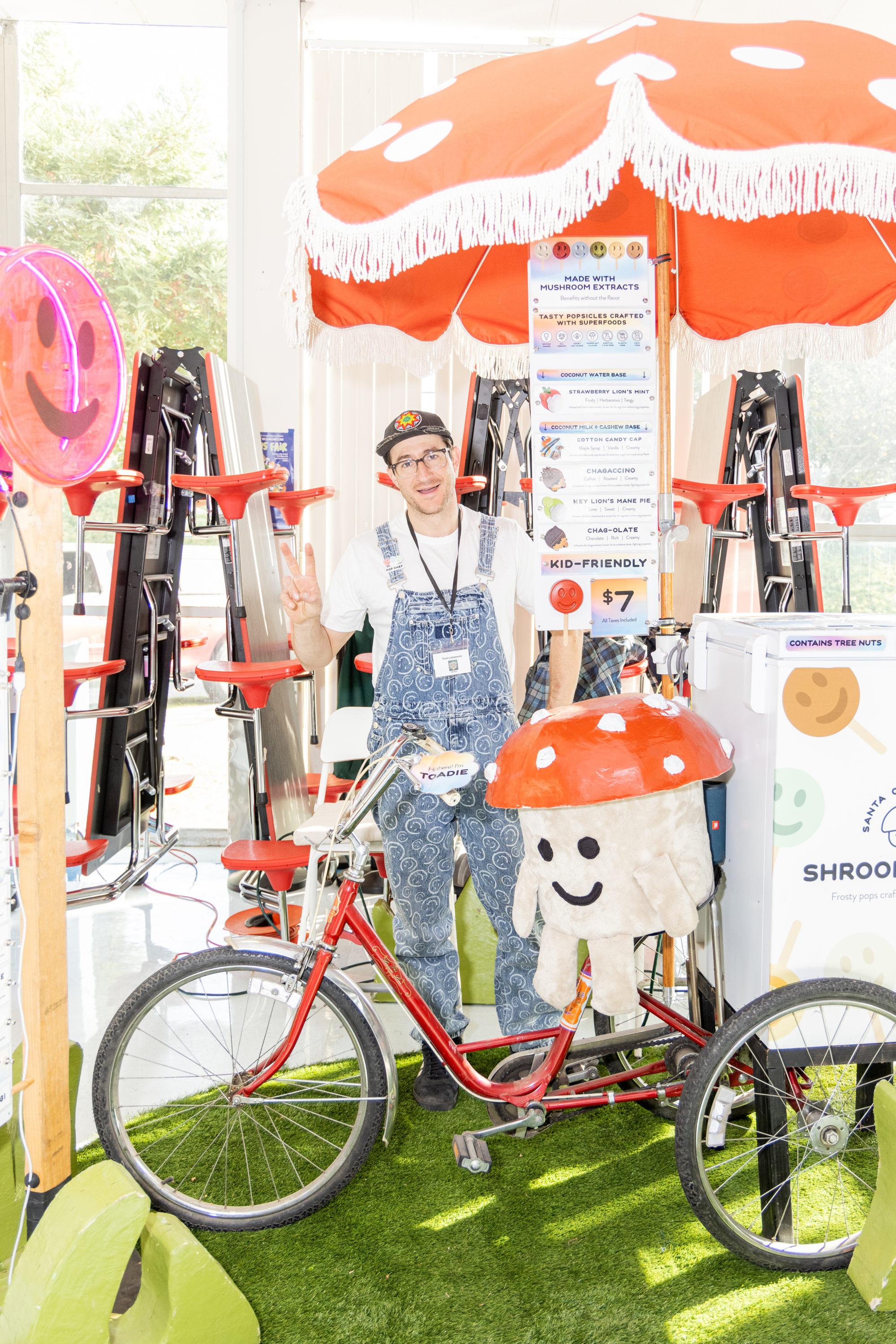 A person in overalls is standing next to a red bicycle under a mushroom-themed umbrella. They're smiling, with a peace sign, surrounded by colorful decor.