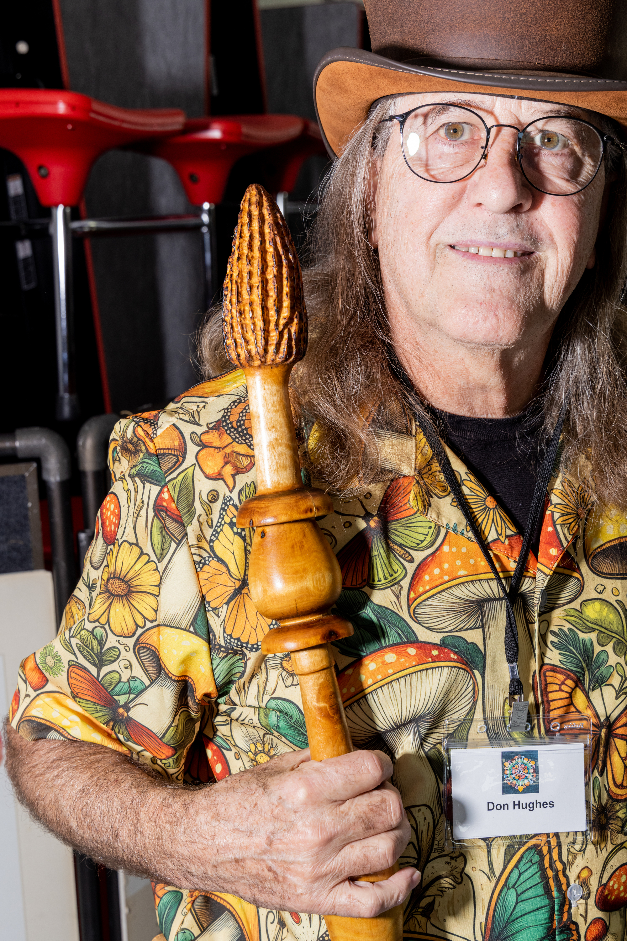 A person with long hair and glasses holds a wooden staff topped with a carved design. They wear a colorful shirt with mushrooms and butterflies.