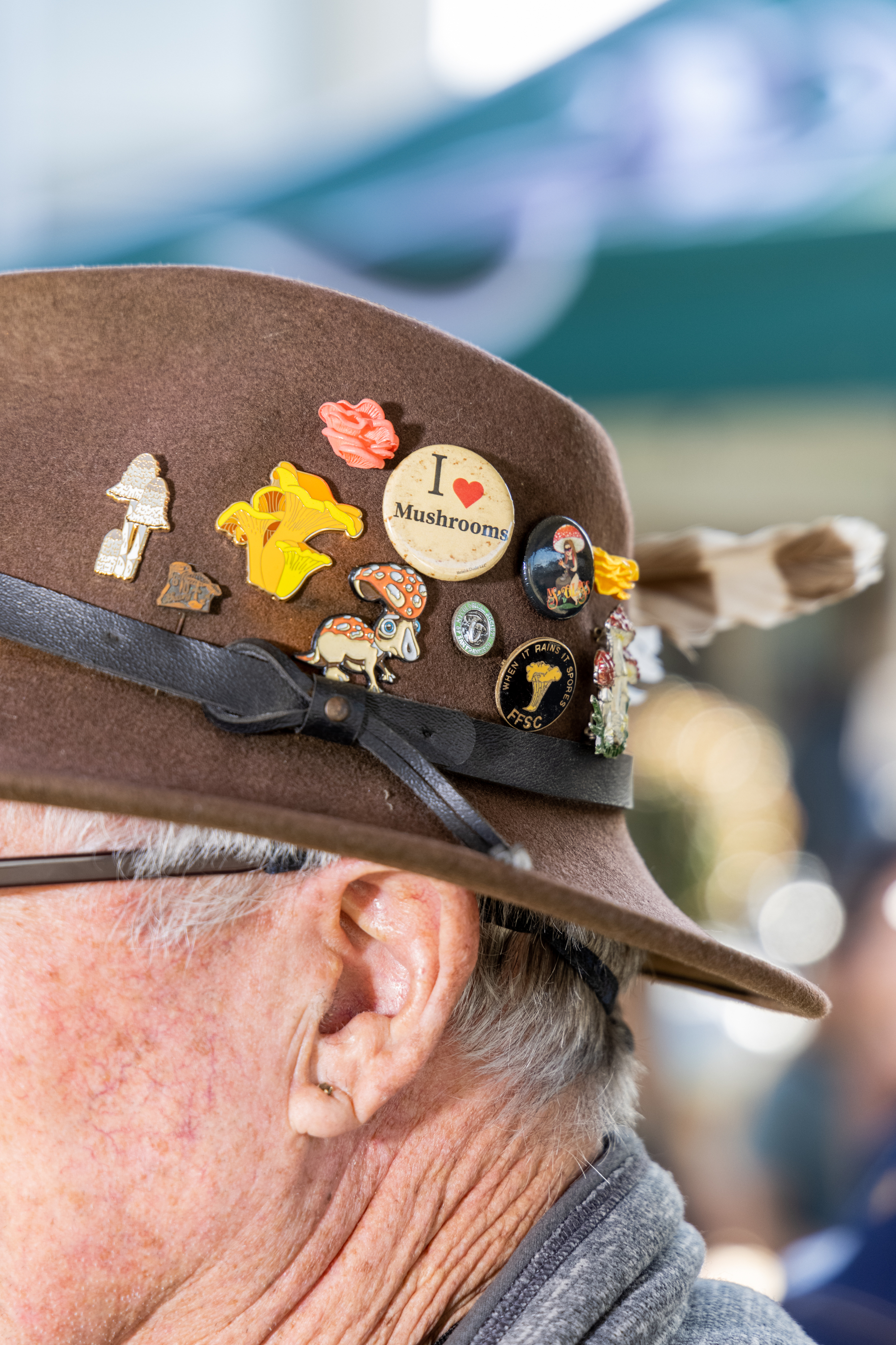 An older man wearing a brown hat adorned with a variety of colorful mushroom pins, including one that says &quot;I love Mushrooms,&quot; and a feather tucked into the band.