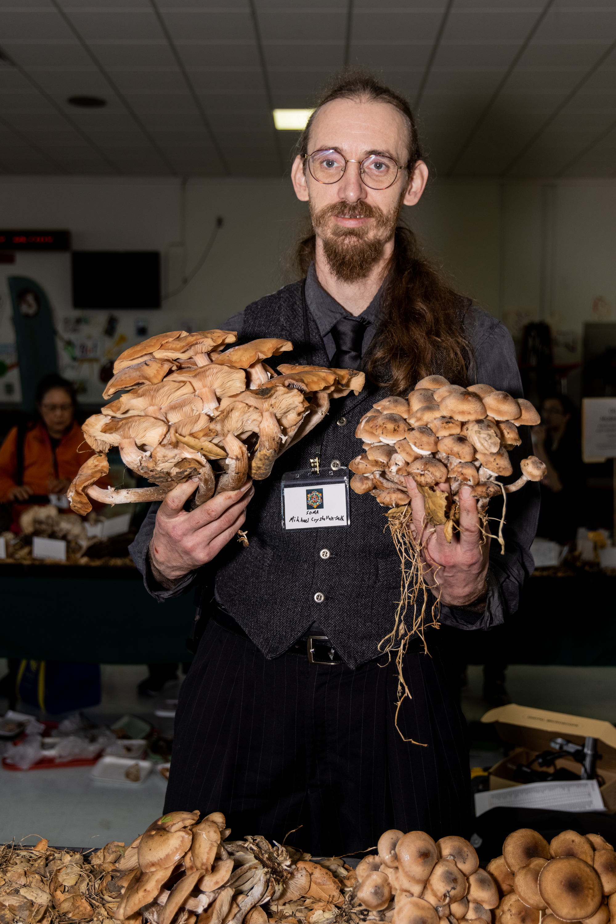 A man with long hair and glasses holds two large clusters of mushrooms, wearing a dark vest and tie. The room in the background has people and tables.