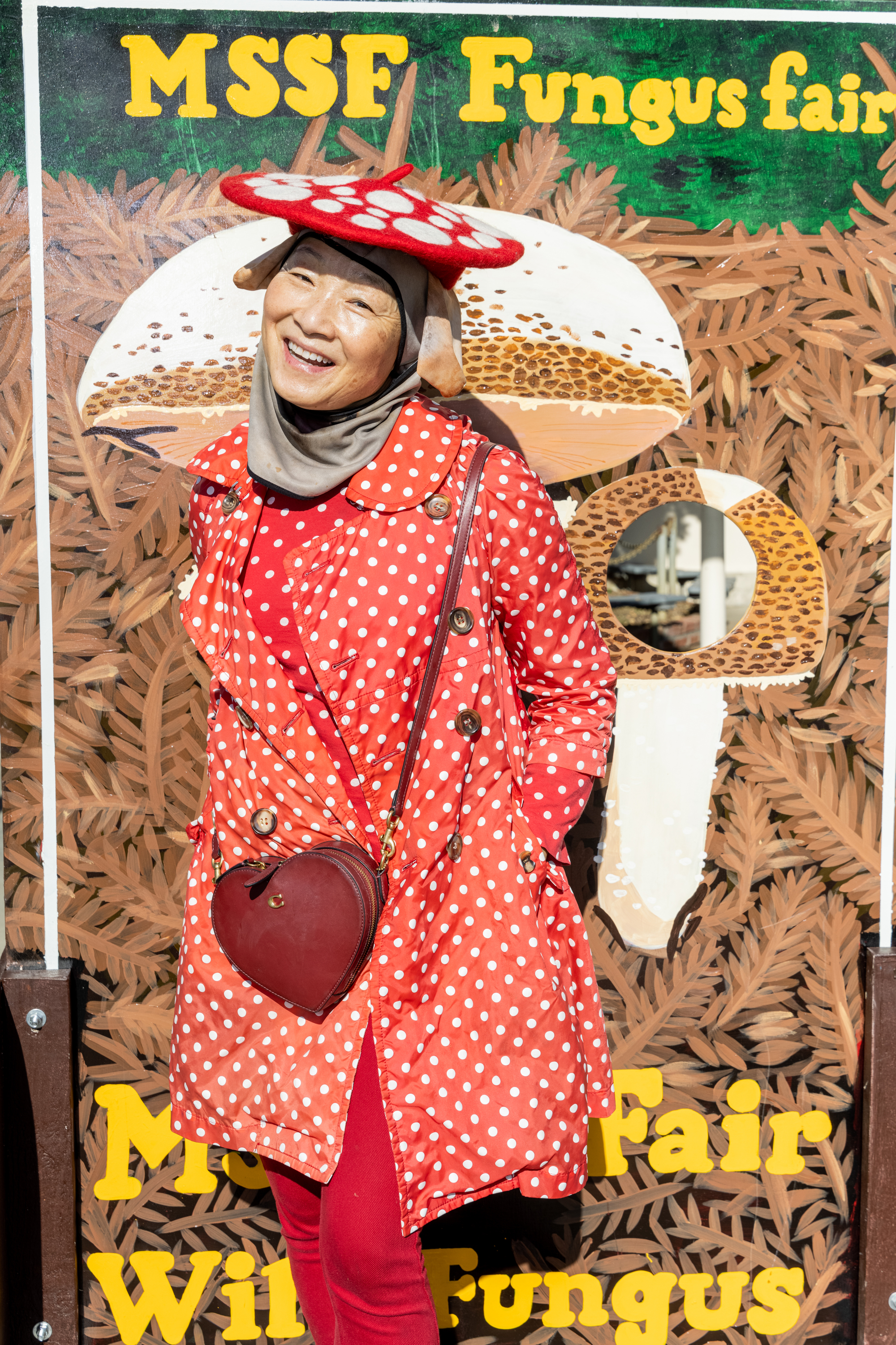 A person smiles in front of a fungus fair sign. They're wearing a red polka-dot outfit and mushroom hat, and have a heart-shaped purse.