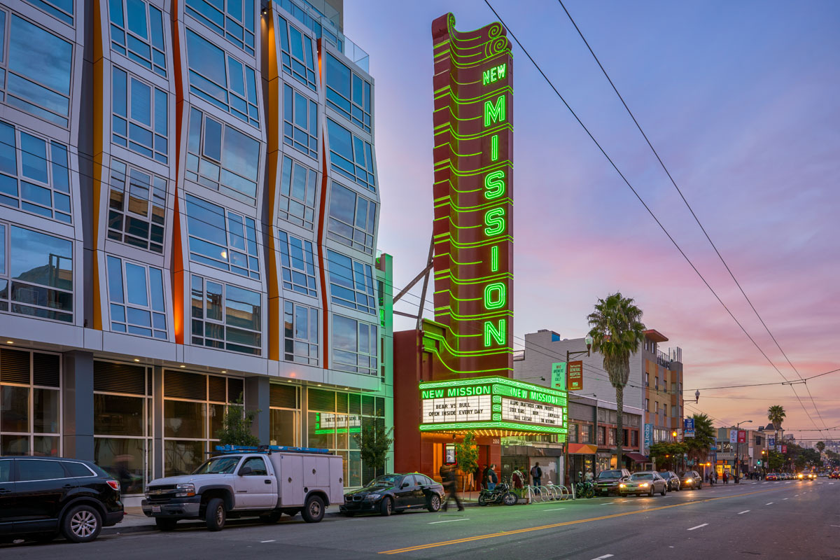 The image shows a street view at sunset with a tall, neon-lit sign reading "New Mission." Modern buildings and parked cars line the road. The sky is pink and purple.