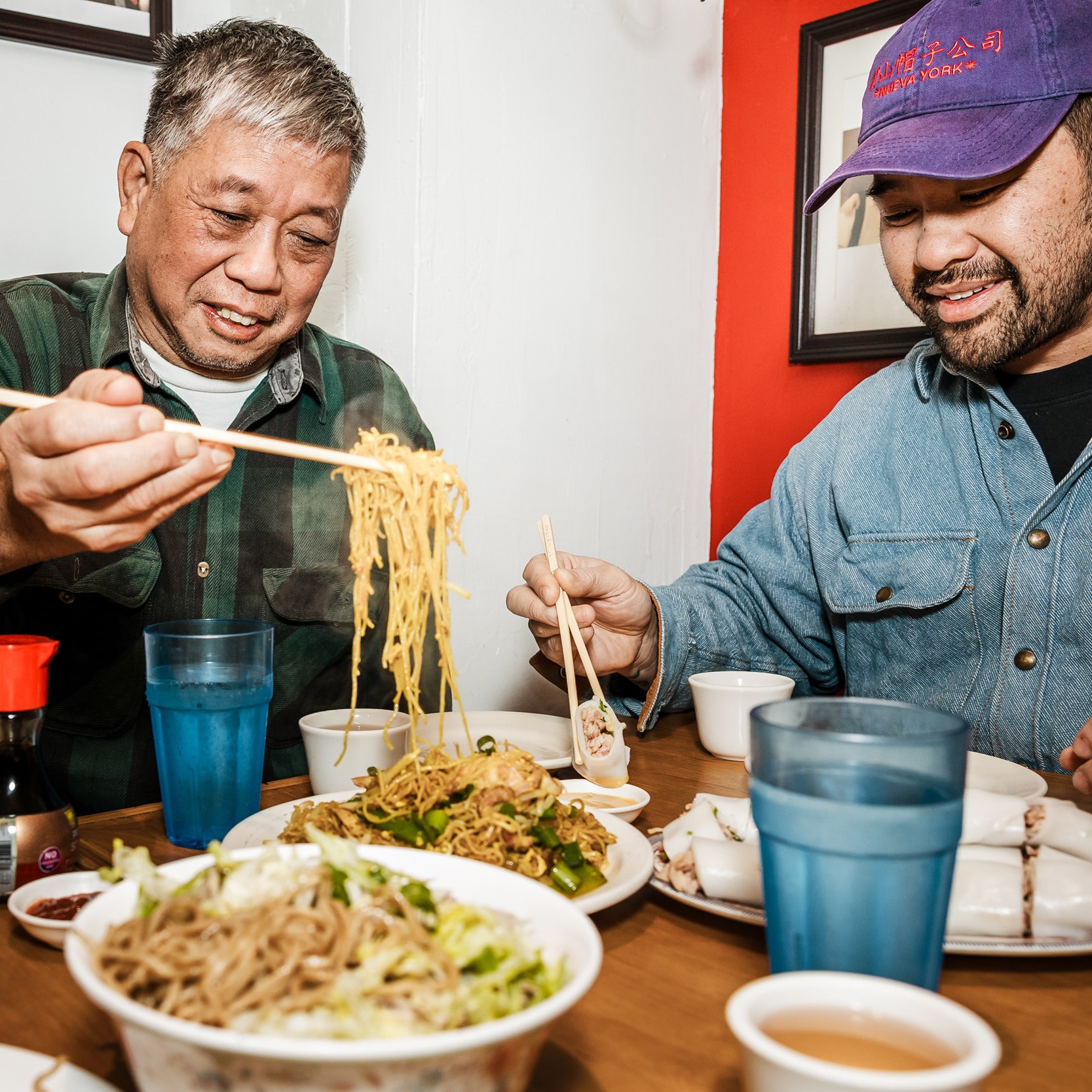 Two men sit at a table filled with Asian dishes, using chopsticks. One man lifts noodles; the other holds a dumpling. Blue cups and a soy sauce bottle are visible.