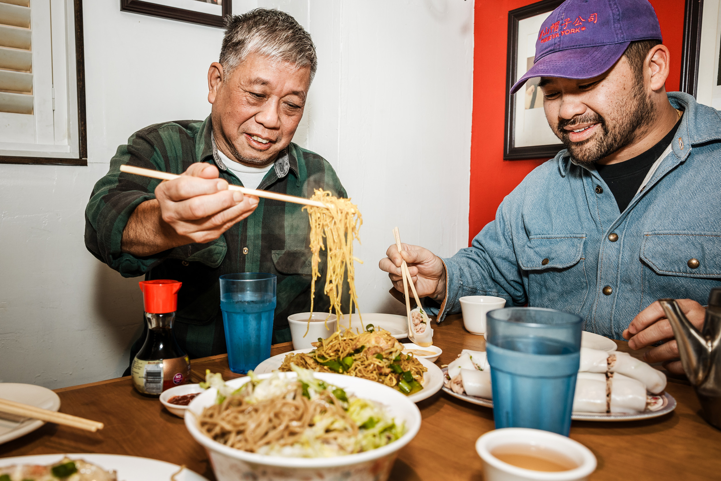 Two men sit at a table filled with Asian dishes, using chopsticks. One man lifts noodles; the other holds a dumpling. Blue cups and a soy sauce bottle are visible.