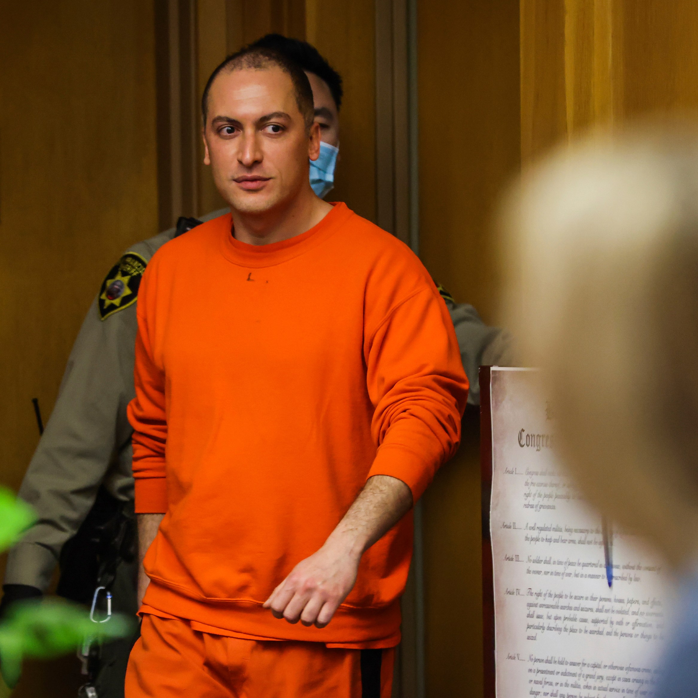 A person in an orange jumpsuit walks in a hallway, accompanied by a law enforcement officer. Papers and a sign are visible on a wall in the background.