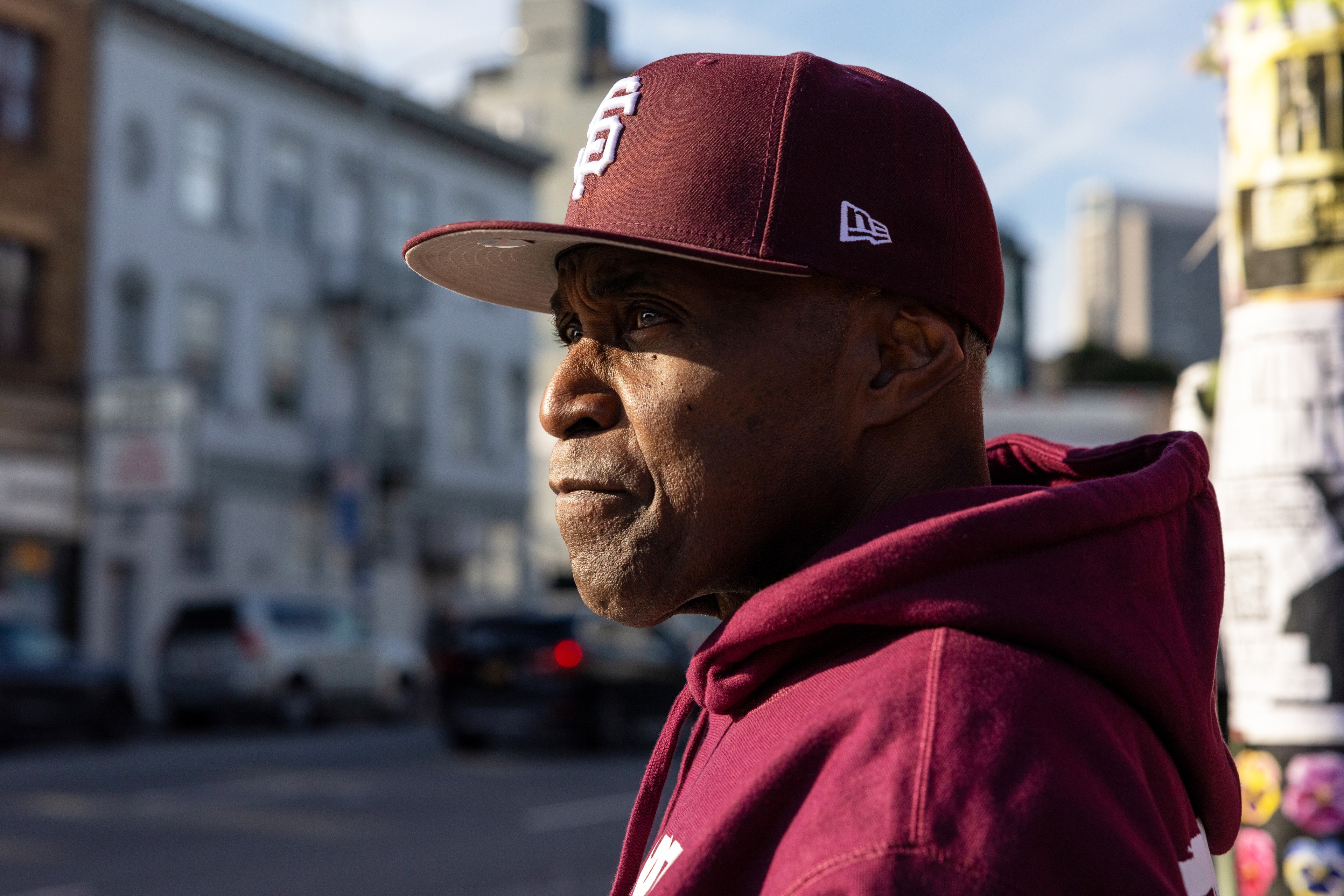 An older man in a burgundy hoodie and cap stands in profile, gazing into the distance on a sunny street with blurred buildings in the background.