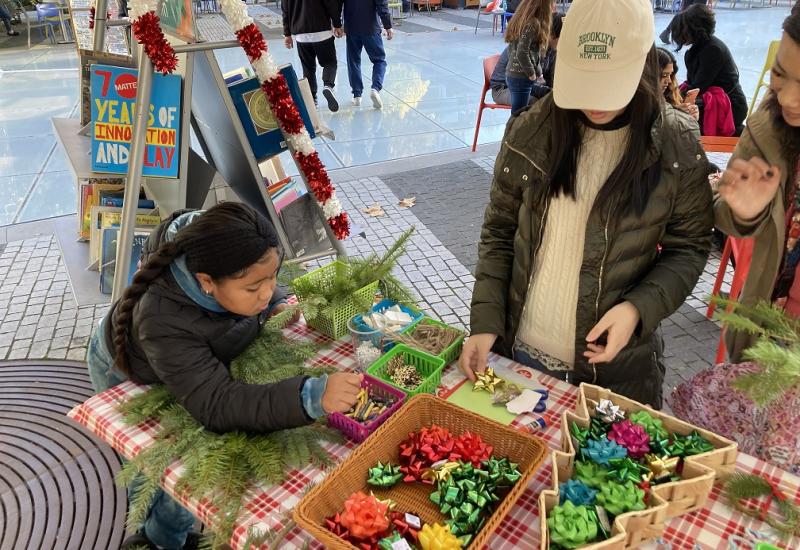 People are making crafts at a table covered with a red-checkered cloth, surrounded by colorful bows, pine branches, and festive decorations.