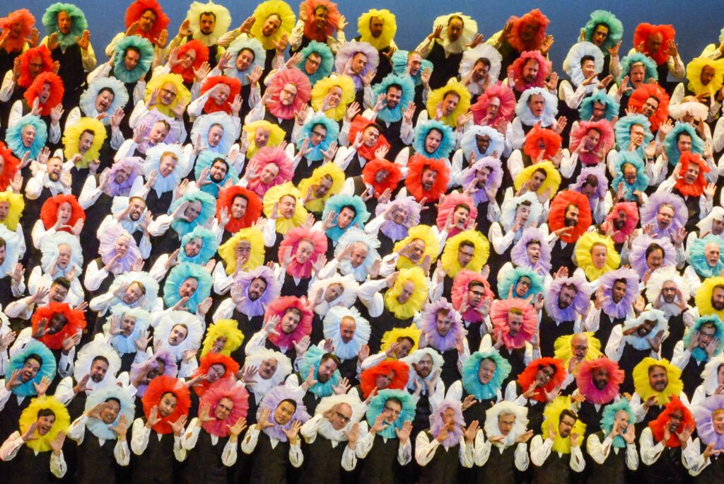 The image shows numerous people wearing colorful frilly collars, arranged in a vibrant, fun pattern. They are smiling and waving, creating a lively scene.