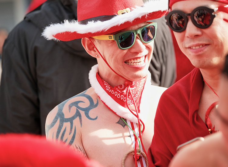 Two smiling individuals in festive attire and sunglasses, one wearing a red cowboy hat and bandana, with tattoos visible, are enjoying a sunny outdoor event.