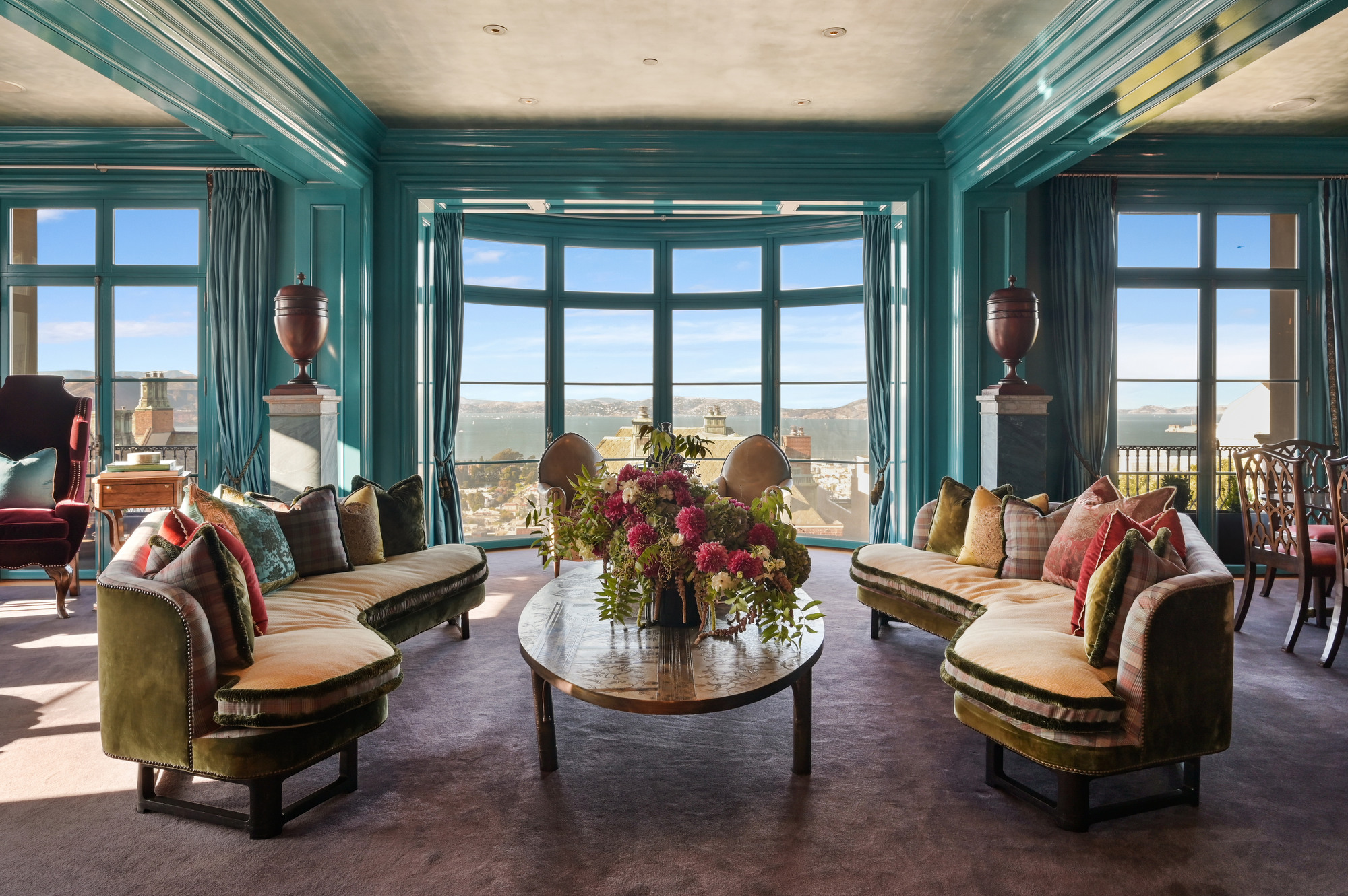 A luxurious living room features velvet sofas, floral arrangements on a central table, and large windows with turquoise frames revealing a scenic outdoor view.