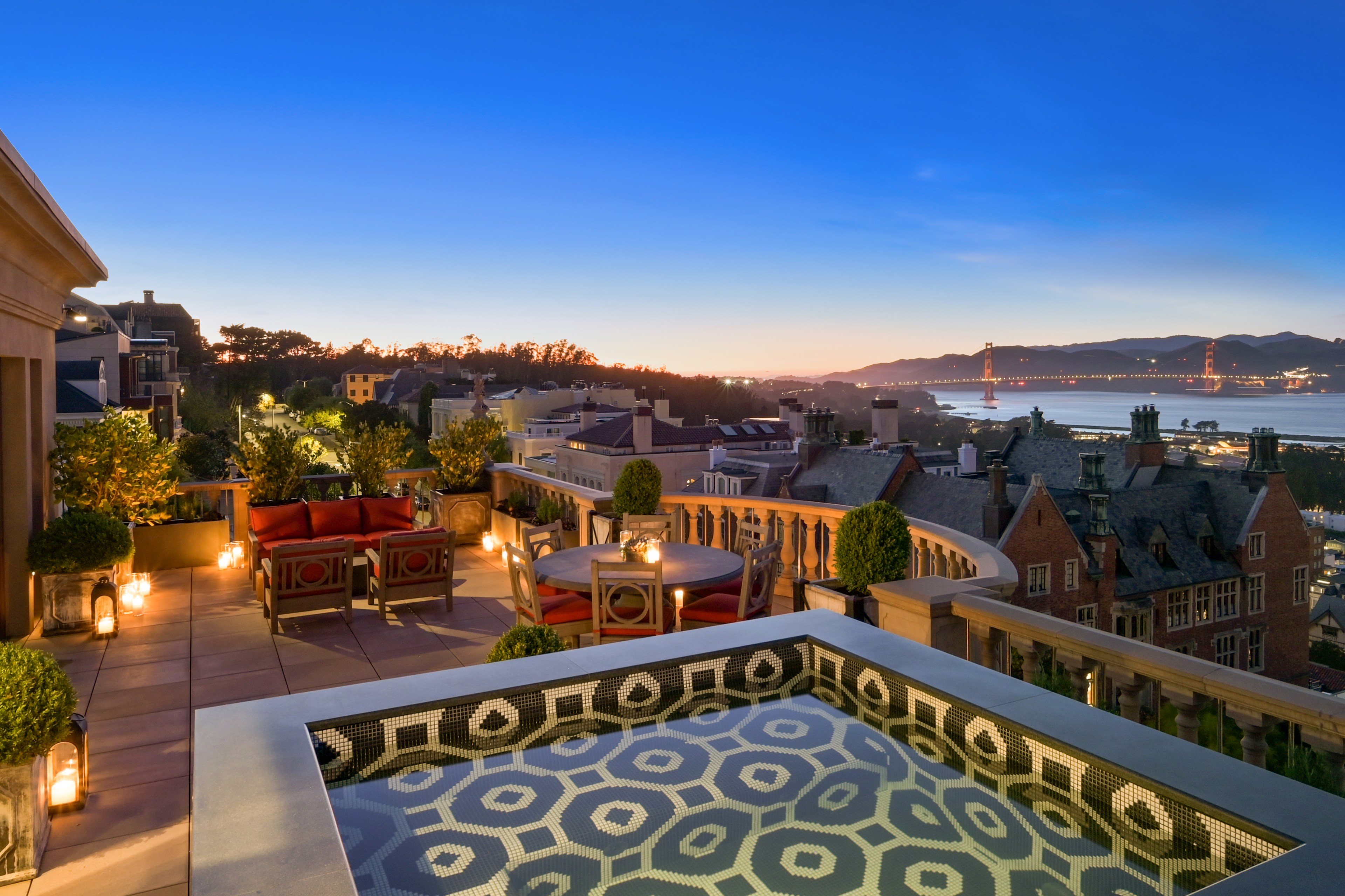 A luxurious rooftop terrace at dusk with seating, decorative lights, and a patterned jacuzzi overlooks a cityscape and the Golden Gate Bridge in the distance.