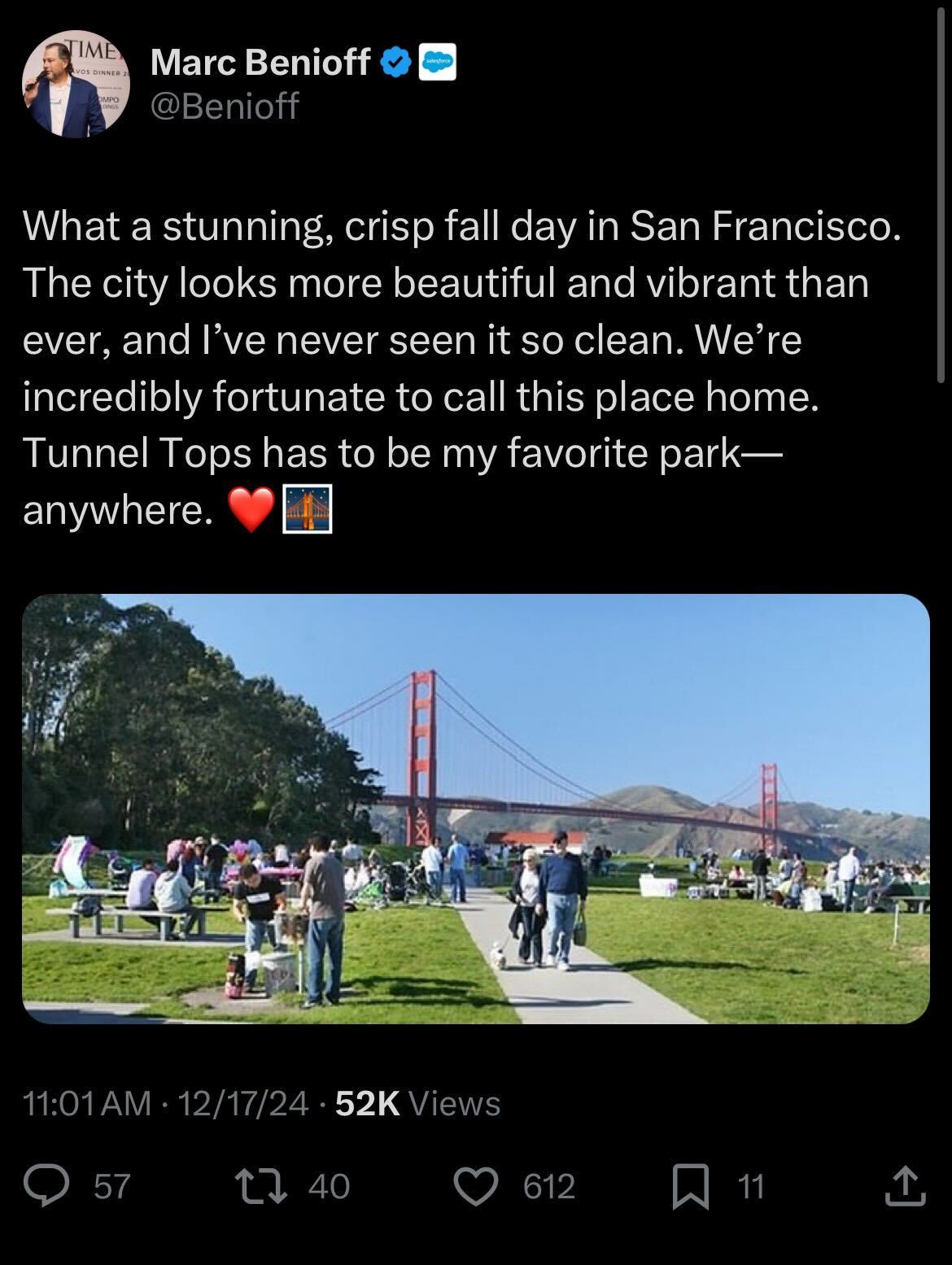 People enjoy a sunny day in a park with picnic tables and paths. The Golden Gate Bridge is visible in the background under a clear blue sky.