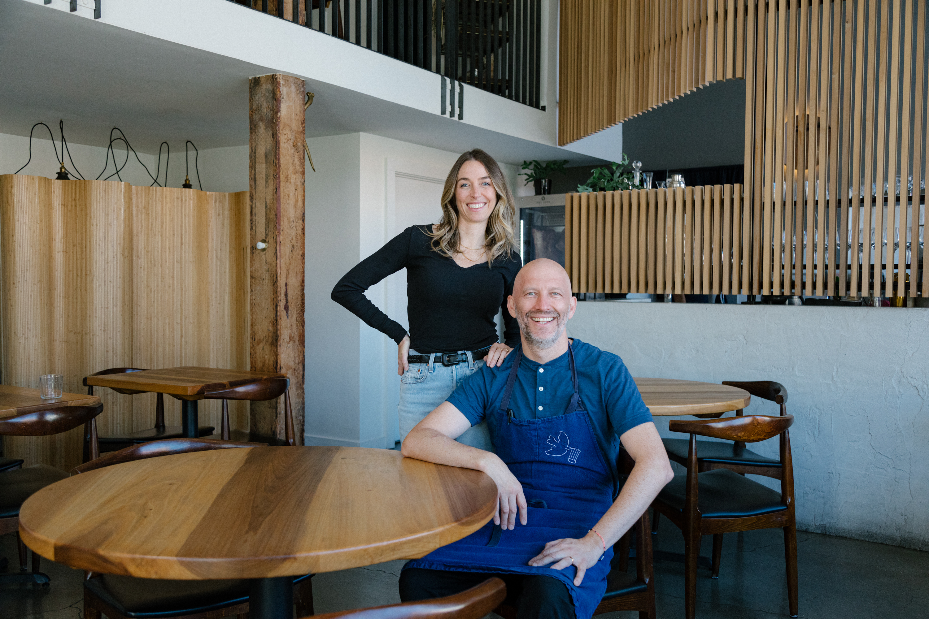 Two people are in a modern restaurant. One is seated at a table, wearing a blue apron, while the other stands beside, smiling. The decor is wooden and minimalist.