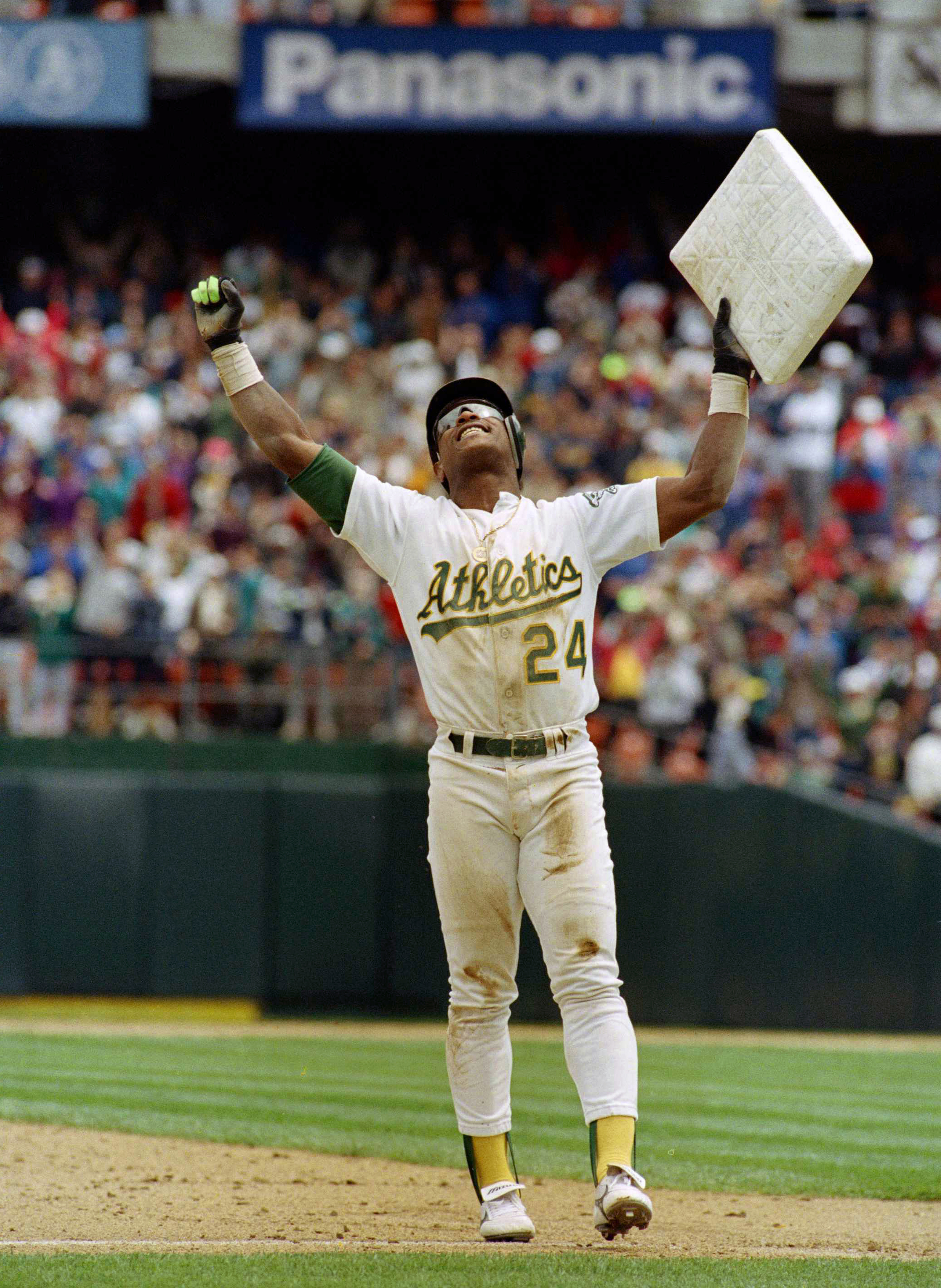 A baseball player in an Athletics uniform, number 24, stands triumphantly holding a base above his head, with a cheering crowd in the background.