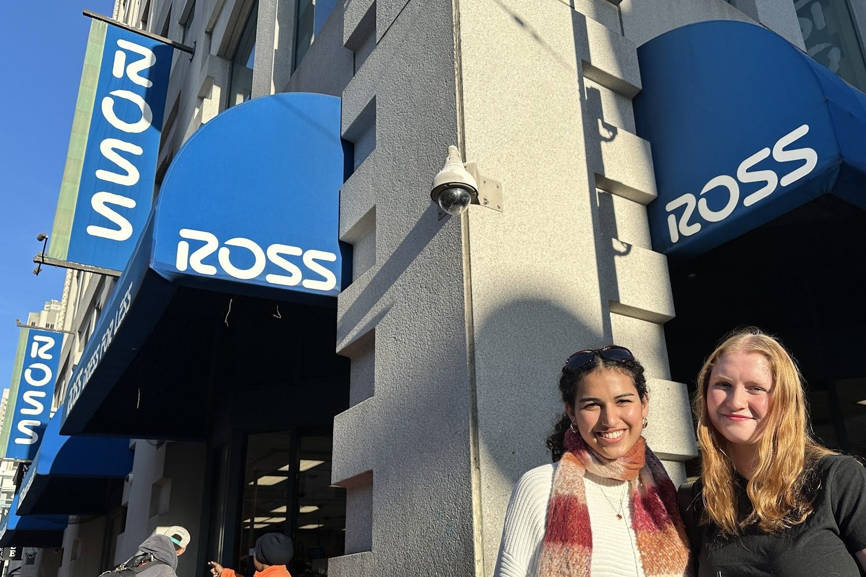 Two people are smiling outside a store with blue awnings displaying the word "ROSS." Another two people are to the left, one pointing towards the store.