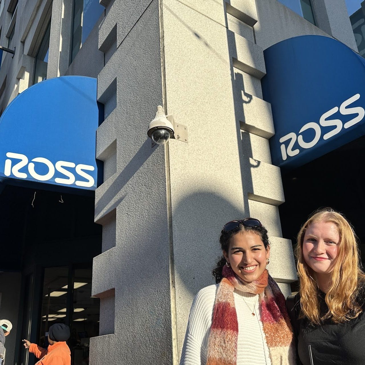 Two people are smiling outside a store with blue awnings displaying the word "ROSS." Another two people are to the left, one pointing towards the store.