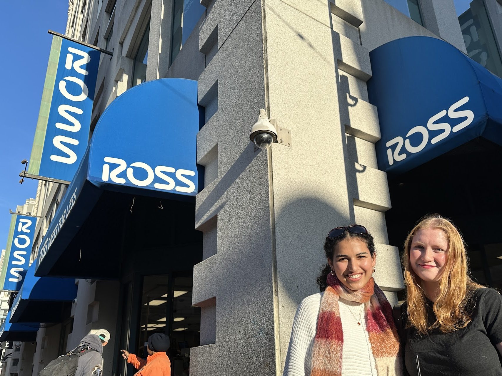 Two people are smiling outside a store with blue awnings displaying the word "ROSS." Another two people are to the left, one pointing towards the store.