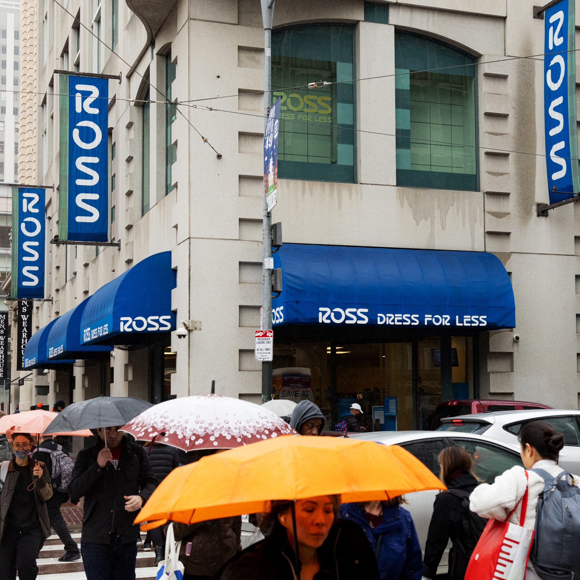 People with umbrellas walk past a &quot;Ross Dress for Less&quot; store on a rainy day. The building has blue awnings and multiple signs displaying the store's name.