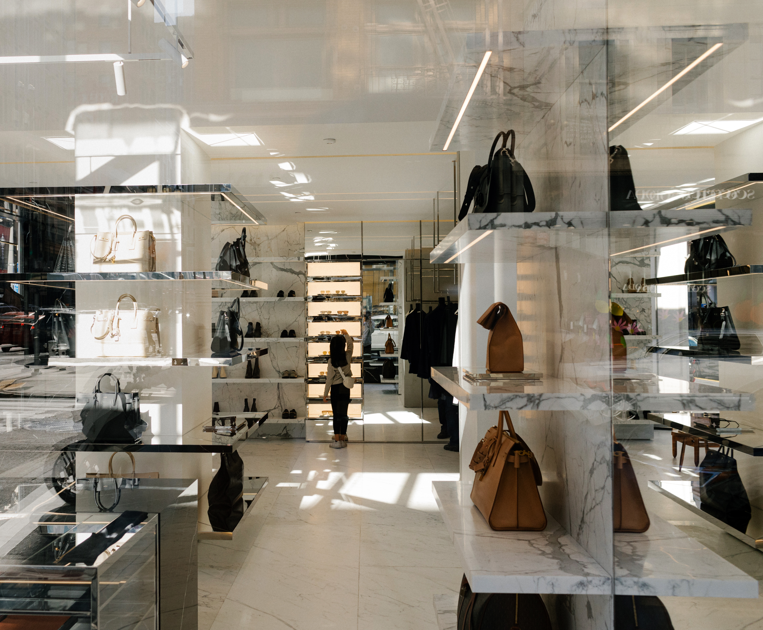 The image shows a luxurious store interior with sleek marble shelves displaying designer handbags and shoes. Sunlight filters through, casting patterns on the floor.