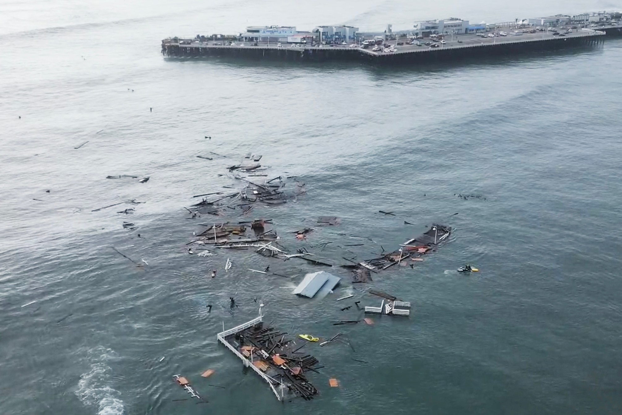 The image shows debris floating in the ocean, with parts of a structure seemingly destroyed near a pier. The water is scattered with various wooden and metal pieces.