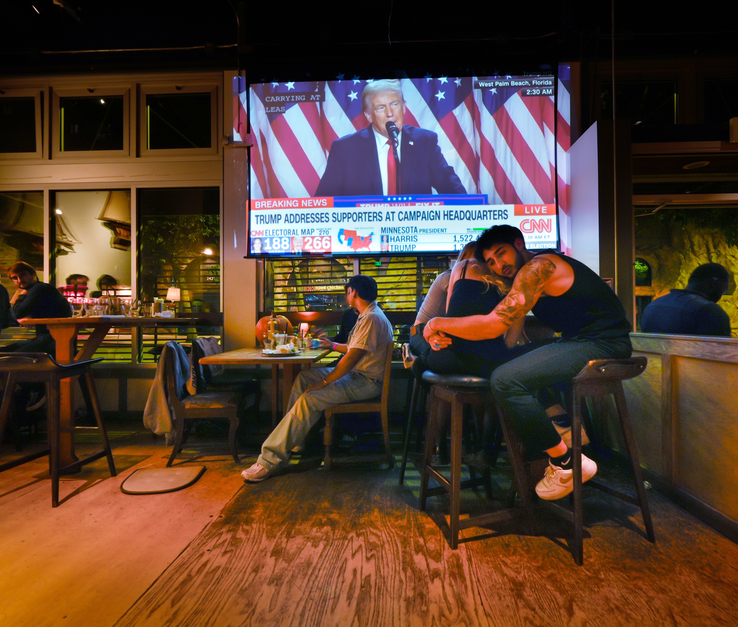 In a dimly lit bar, people watch a TV showing Trump speaking at campaign headquarters, with election results displayed. A couple embraces at the bar.