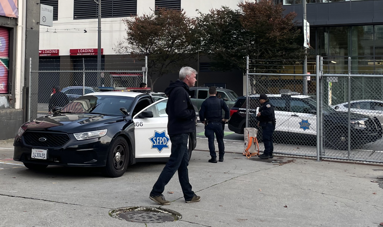 A police car is parked on the street with &quot;SFPD&quot; on the door. Two officers and a man are nearby, standing near a chain-link fence with some trees in the background.