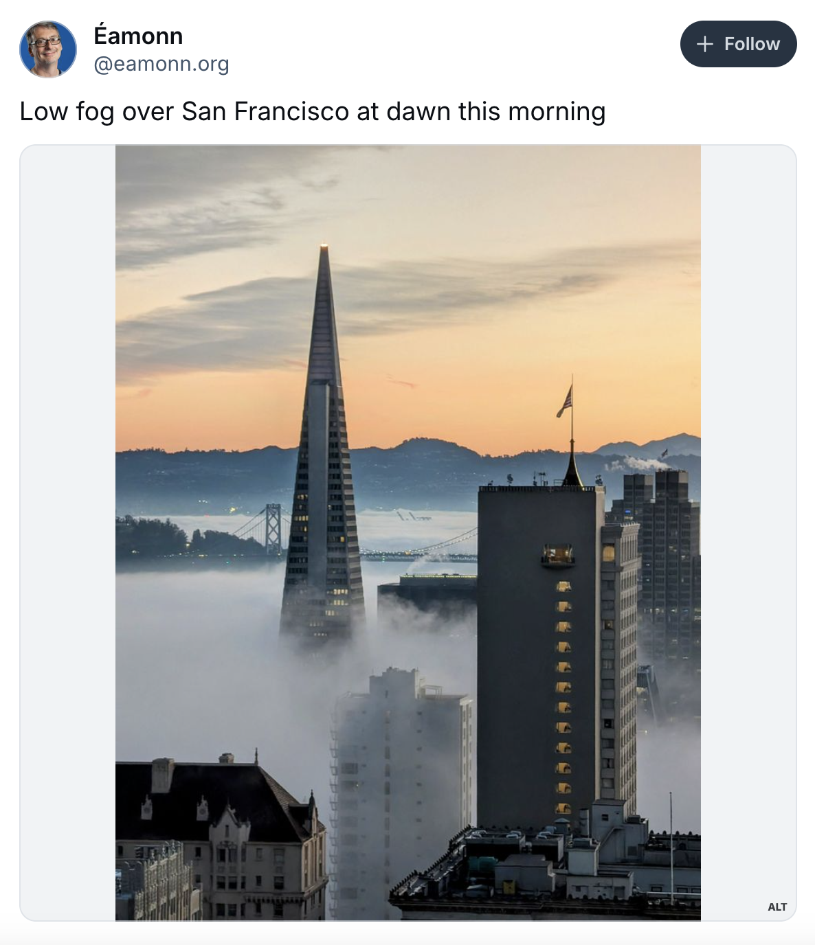 The image shows a foggy San Francisco skyline at dawn, with the Transamerica Pyramid emerging above the fog and a soft, colorful sunrise in the background.