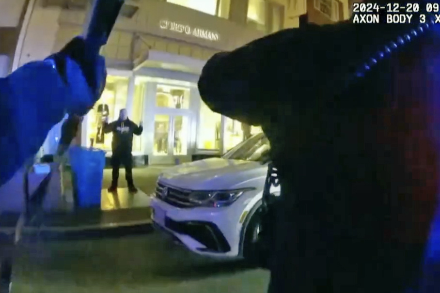 The image shows a nighttime street scene with a white SUV parked in front of a brightly lit storefront. A person stands with arms raised, while police officers are visible in the foreground.