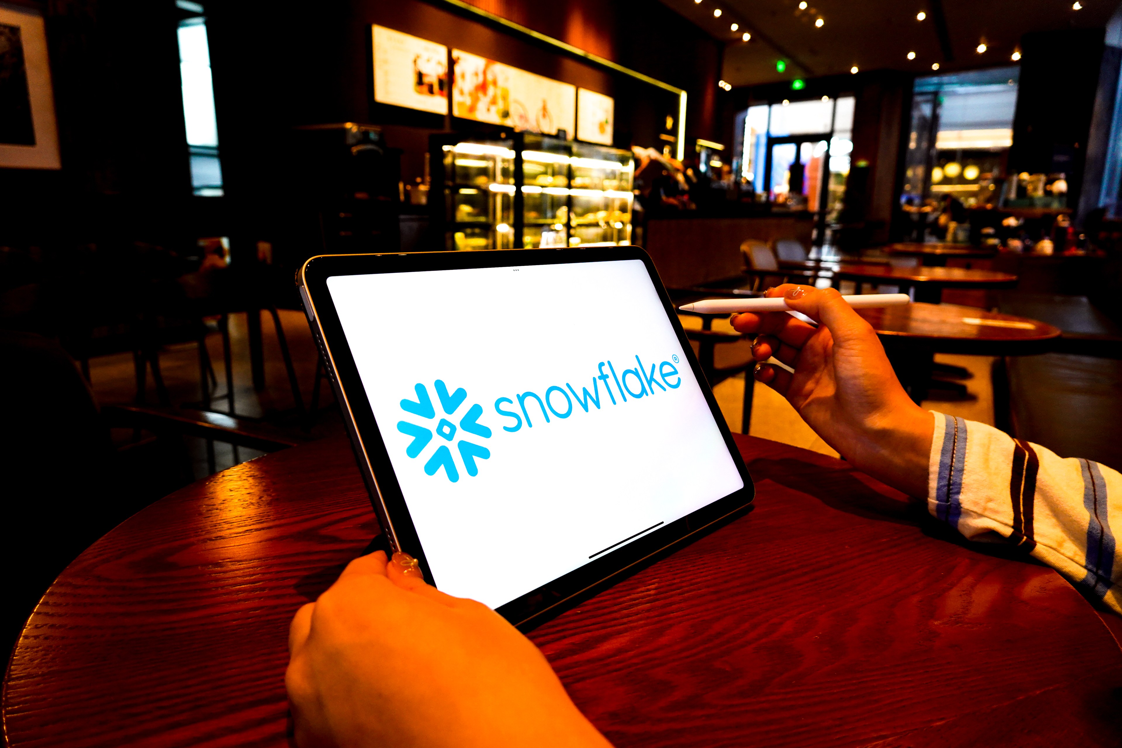 A person holds a tablet displaying the Snowflake logo, seated at a wooden table in a cozy, well-lit cafe with a modern interior.