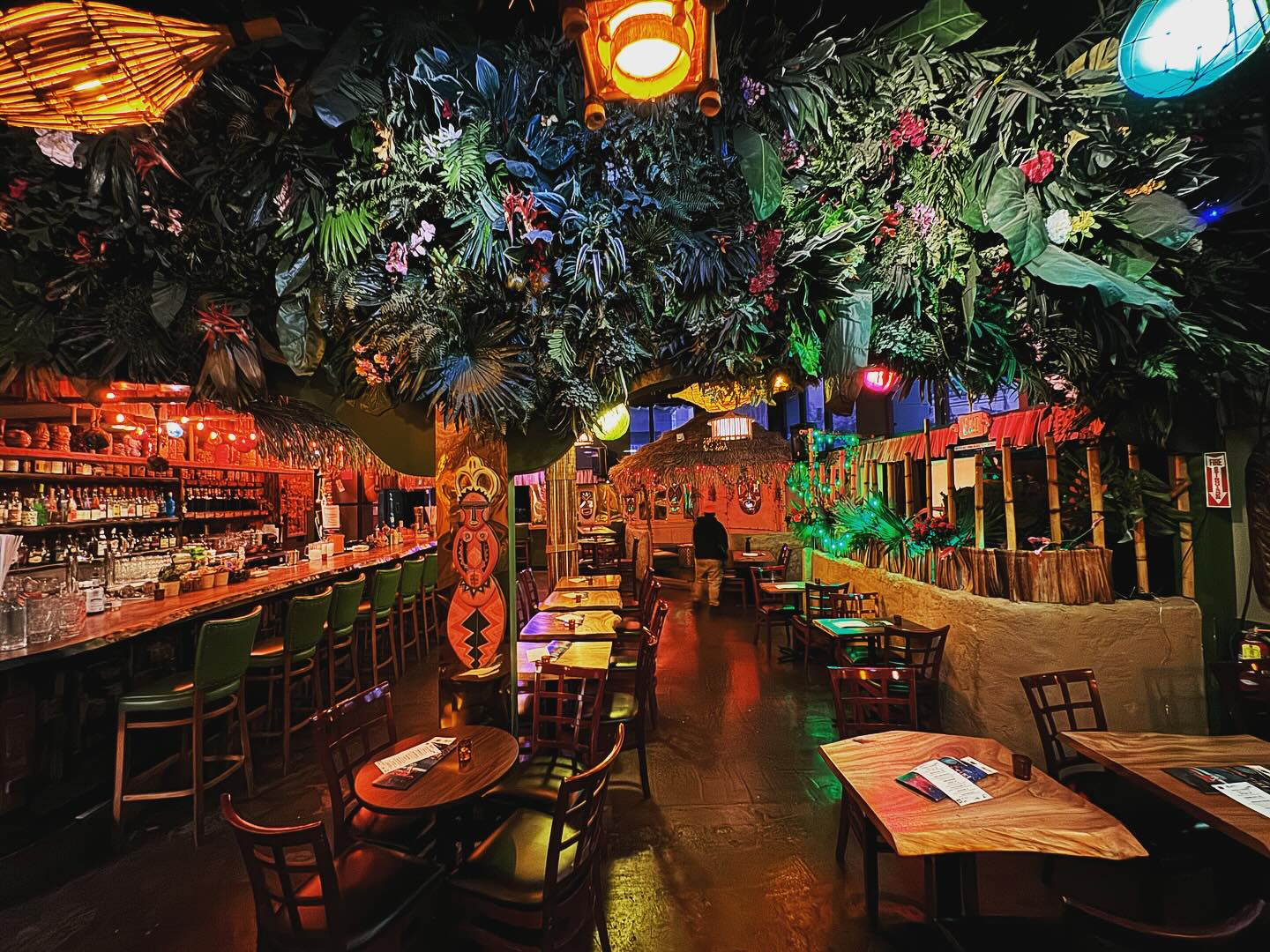 A vibrant tiki bar with a lush, tropical ceiling decor of plants and flowers. There are wooden tables and chairs, bamboo details, and colorful lighting.