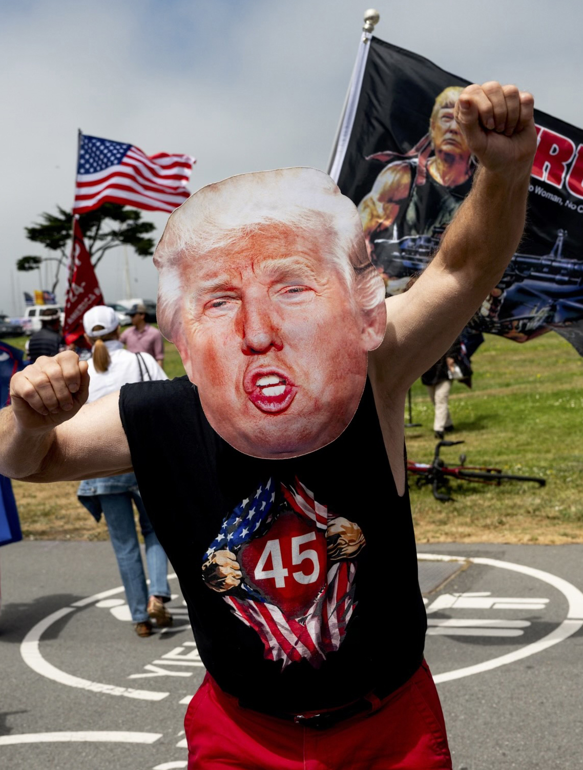 A person wearing a Donald Trump mask and a &quot;45&quot; shirt is posing with clenched fists. American flags are in the background, suggesting a political gathering.