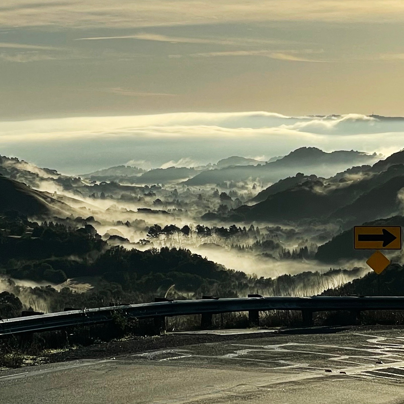 A scenic view of valleys shrouded in mist with rolling hills and a winding road in the foreground, featuring a yellow road sign with a right turn arrow.