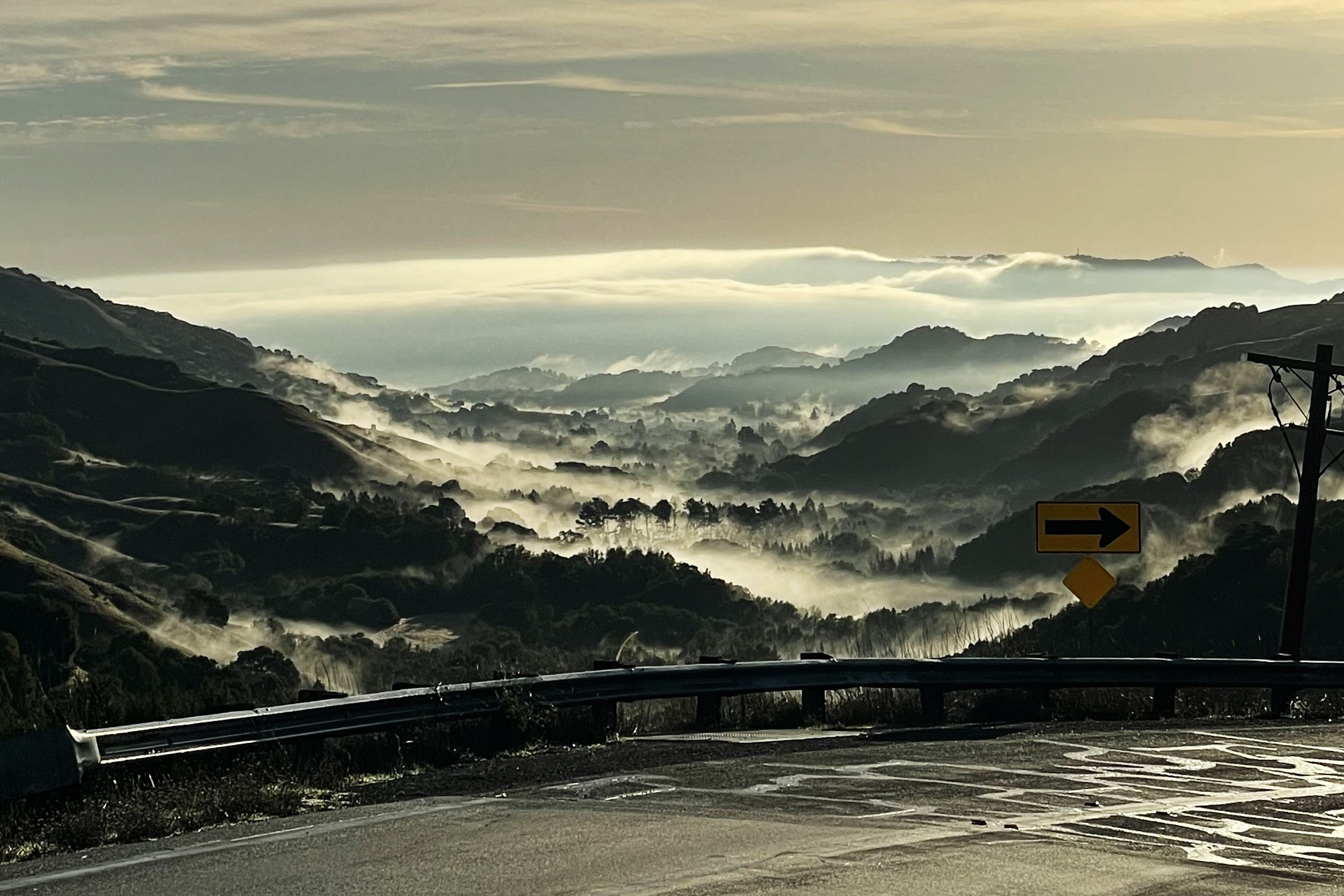 A scenic view of valleys shrouded in mist with rolling hills and a winding road in the foreground, featuring a yellow road sign with a right turn arrow.