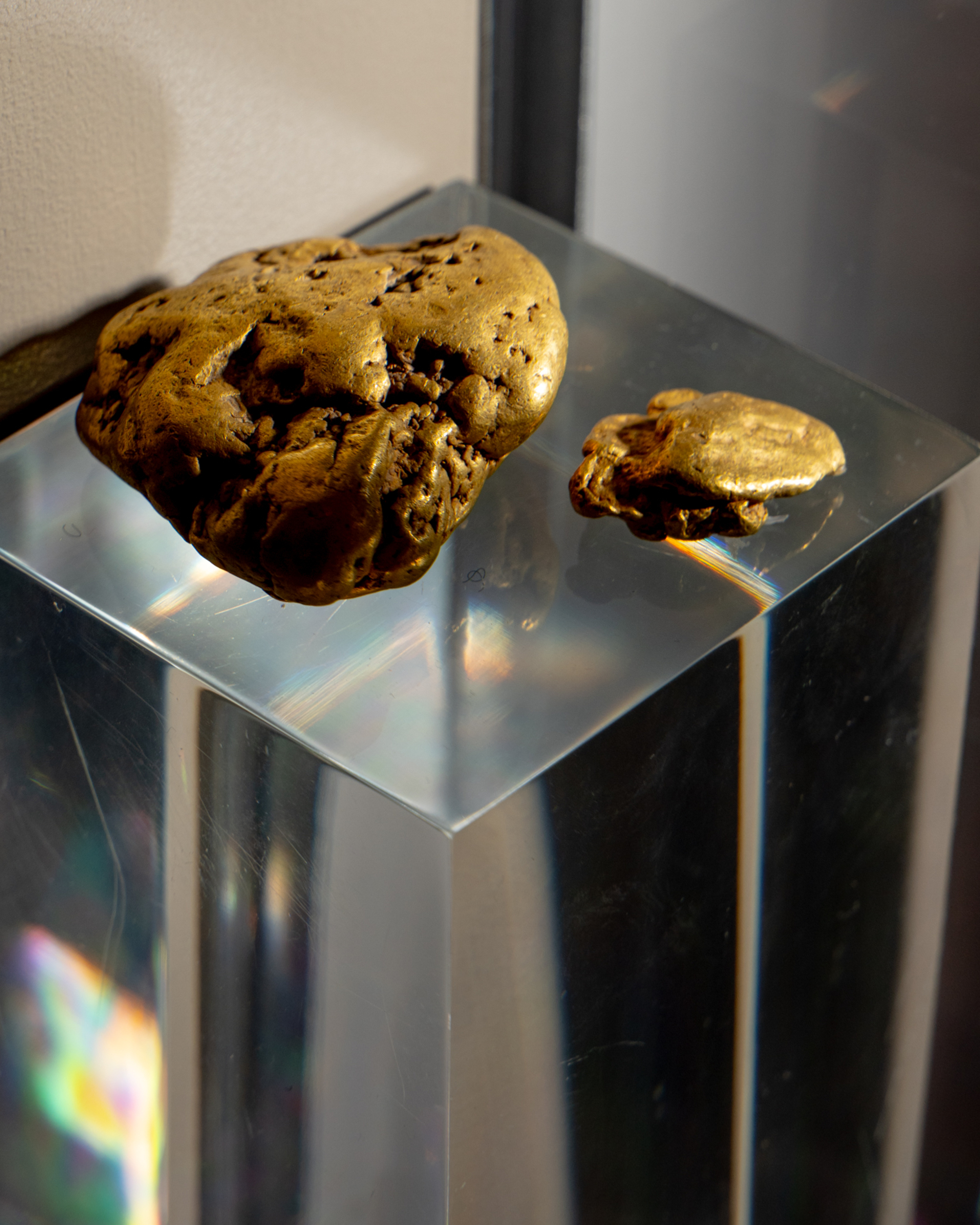 Two irregularly shaped gold nuggets rest on a clear, reflective display stand, with a beige background and faint light reflections visible on the surface.