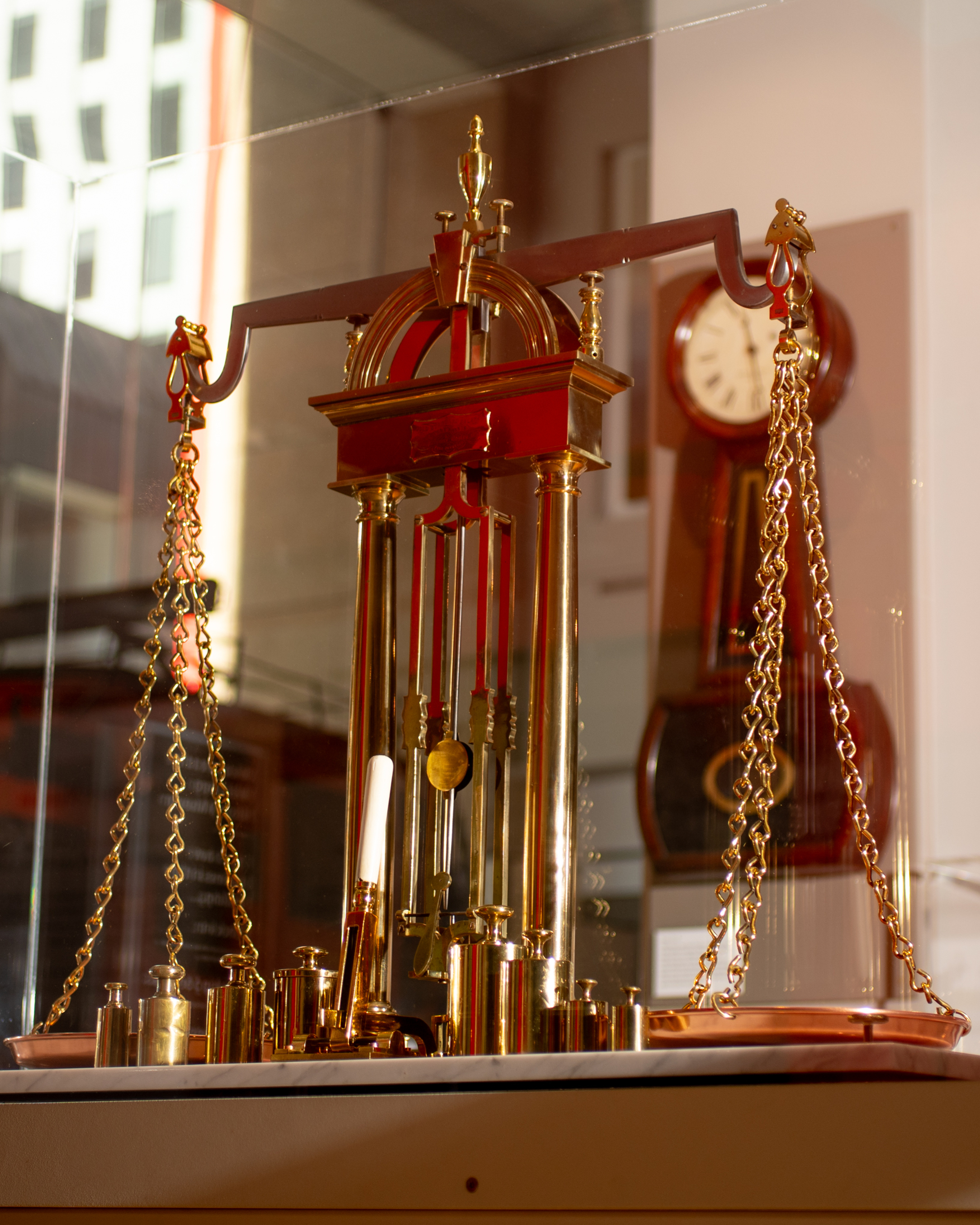 The image shows a decorative brass scale with chains and weights, set on a display. In the background, there's a clock on the wall.